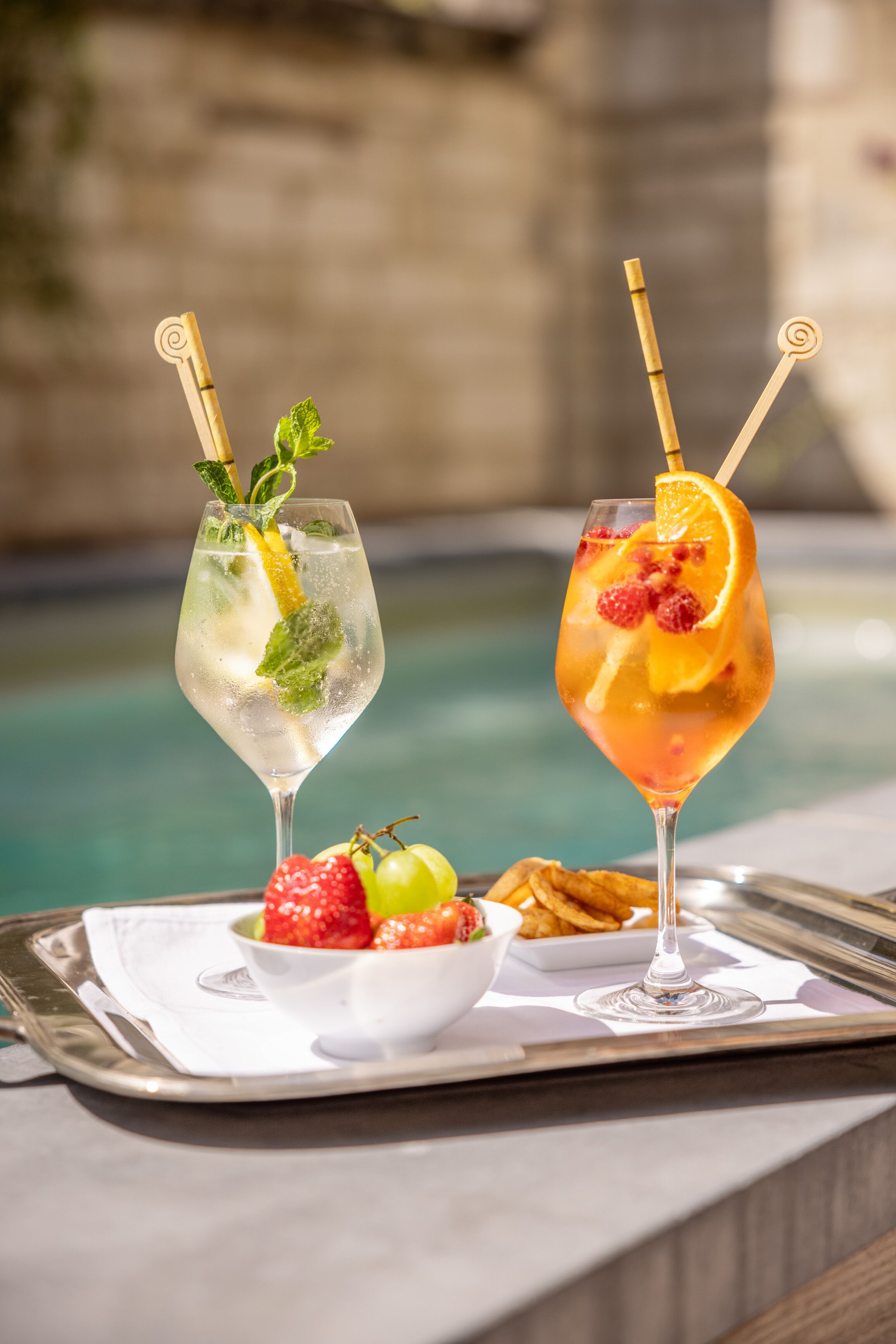 Deux cocktails servis sur un plateau, accompagnés de fraises et d’un saladier de fruits, près d’une piscine.
