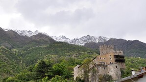 Un edificio histórico de piedra con una torre, rodeado de exuberantes colinas verdes y montañas nevadas al fondo.
