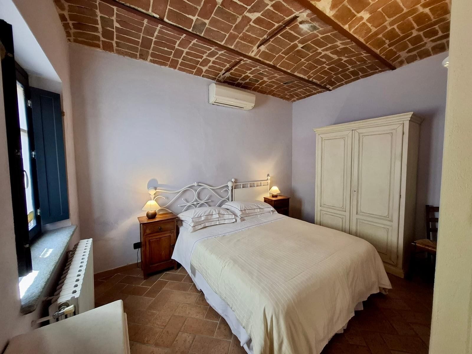 A bedroom with a brick ceiling, a white bed, two bedside tables with lamps, a white wardrobe, and a radiator.
