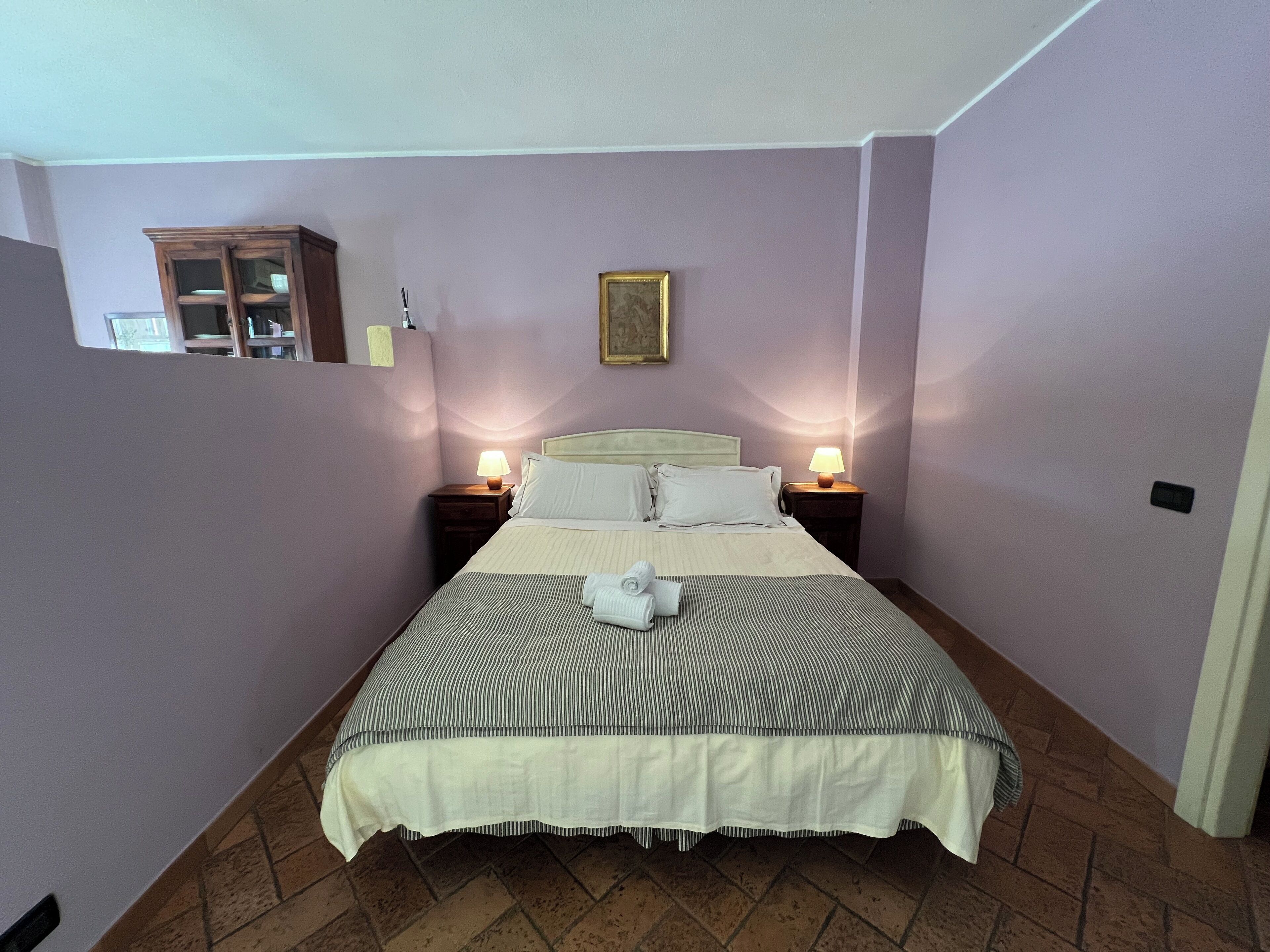 Cama bem arrumada com colcha listrada e almofadas, duas luminárias de cabeceira, um pequeno armário de madeira e um quadro na parede.