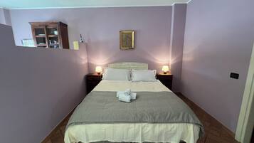 Cama bem arrumada com colcha listrada e almofadas, duas luminárias de cabeceira, um pequeno armário de madeira e um quadro na parede.