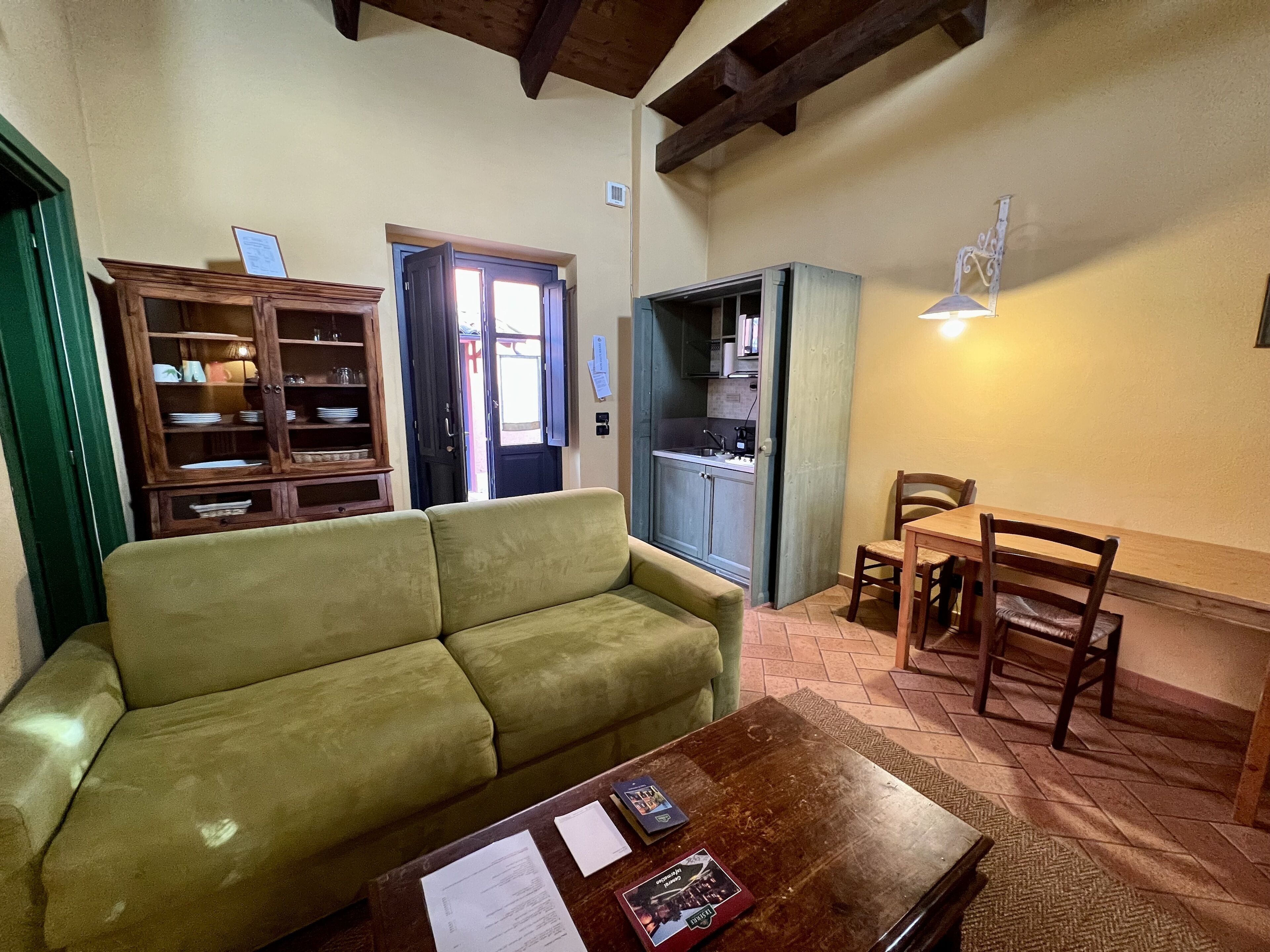 A cozy living room with a green sofa, a wooden coffee table with documents, a dining area with a wooden table and chairs, and a kitchen area with cabinets and a window.