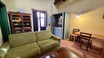 A cozy living room with a green sofa, a wooden coffee table with documents, a dining area with a wooden table and chairs, and a kitchen area with cabinets and a window.