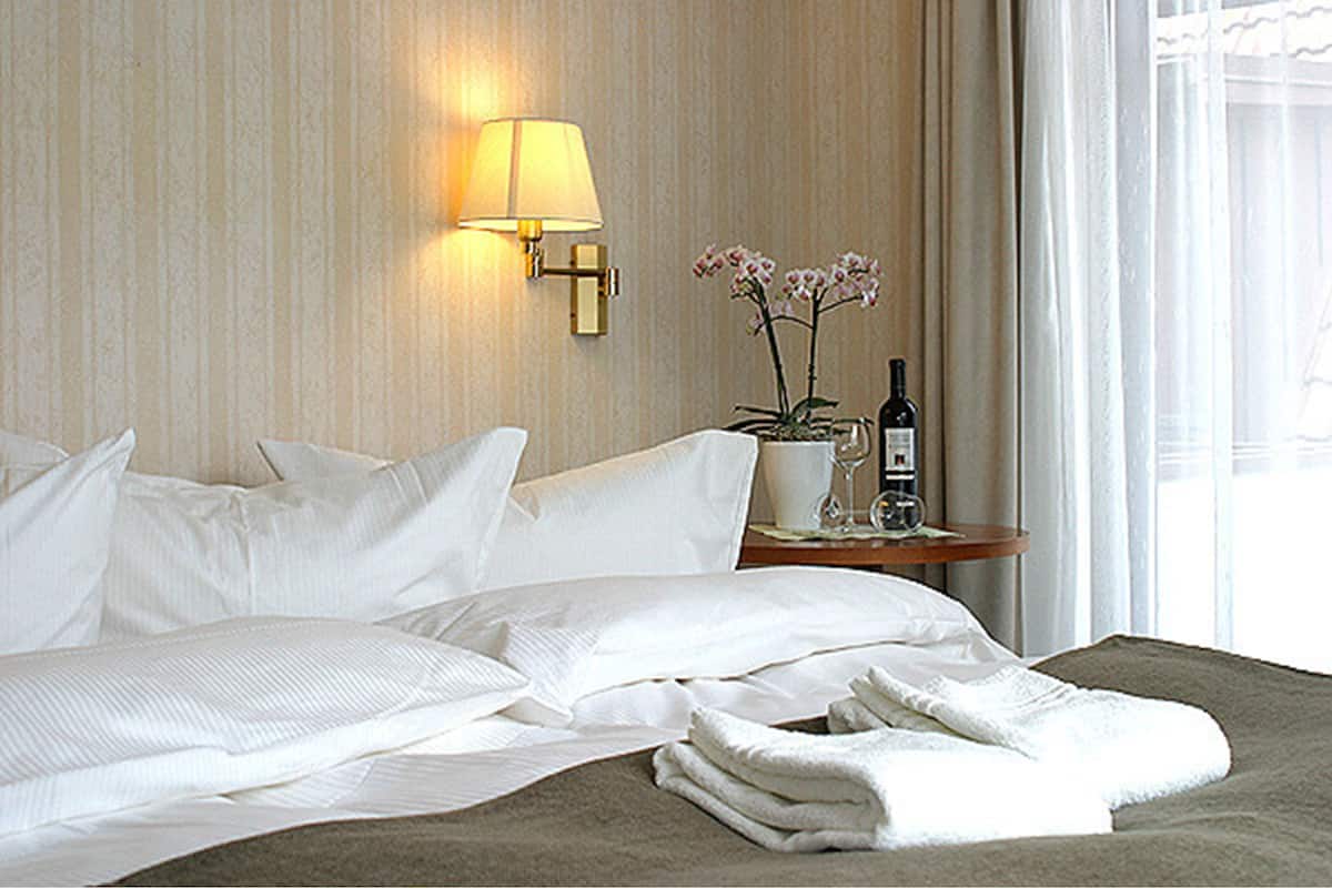 Un lit bien fait, avec du linge de lit blanc, une lampe de chevet, une petite table sur laquelle se trouvent une bouteille de vin et un verre, et une orchidée en pot.
