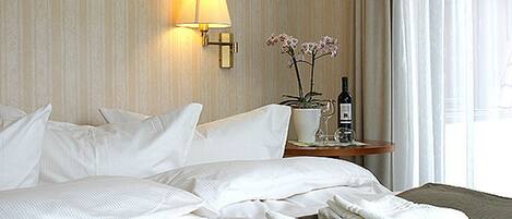 A neatly made bed with white linens, a bedside lamp, a small table with a wine bottle and a glass, and a potted orchid.