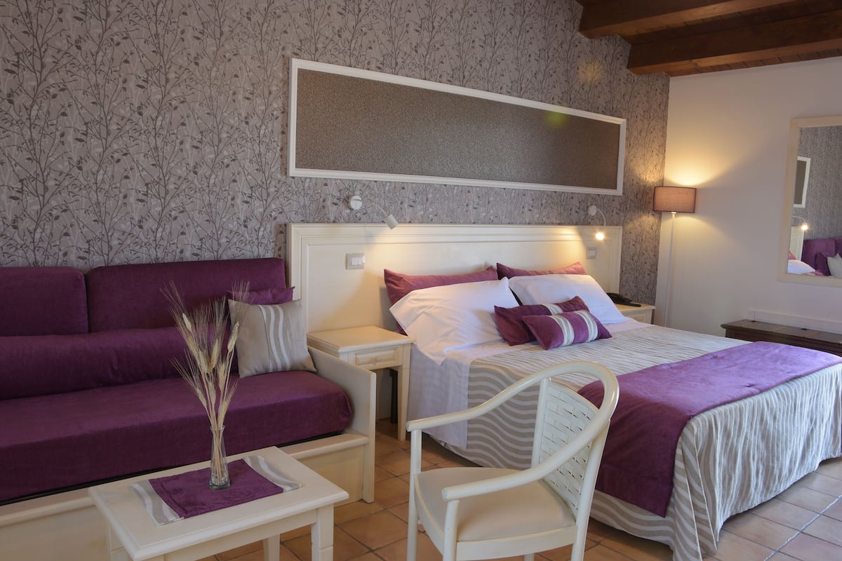 A bedroom with a bed, a sofa, a chair, a nightstand, and a bedside table.