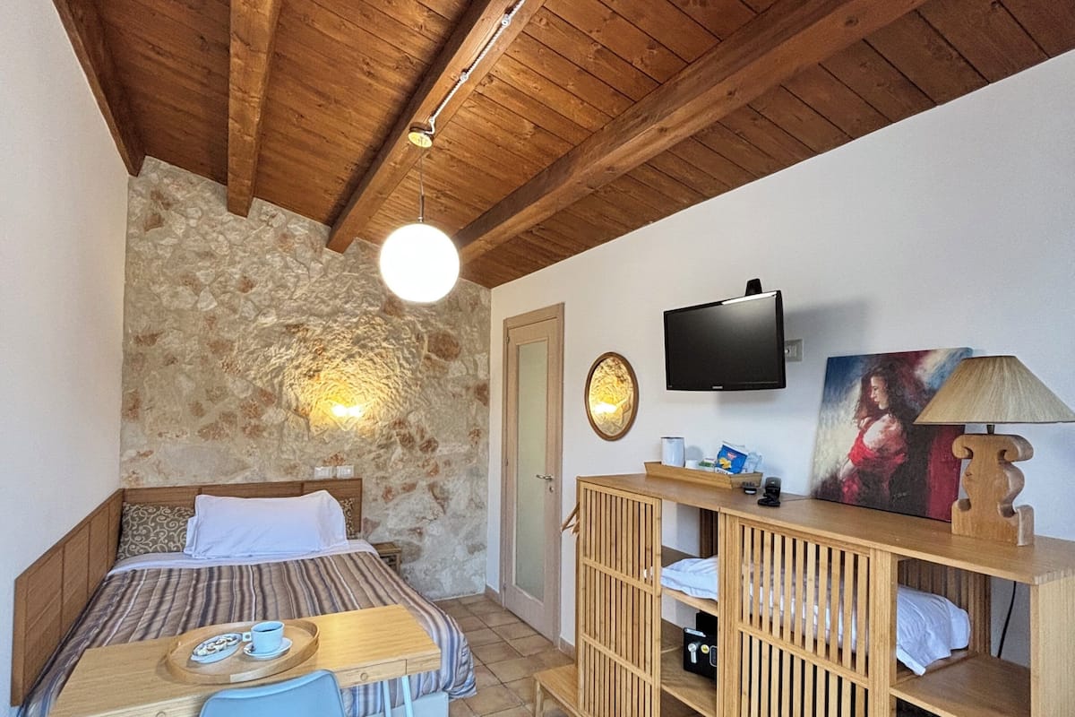 A bedroom with a bed, a desk, a chair, a TV, and a stone wall.