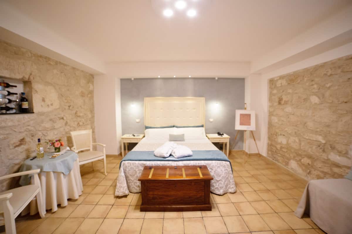 A bedroom with a large bed, a wooden bench, a small table with a tablecloth, and a chair.
