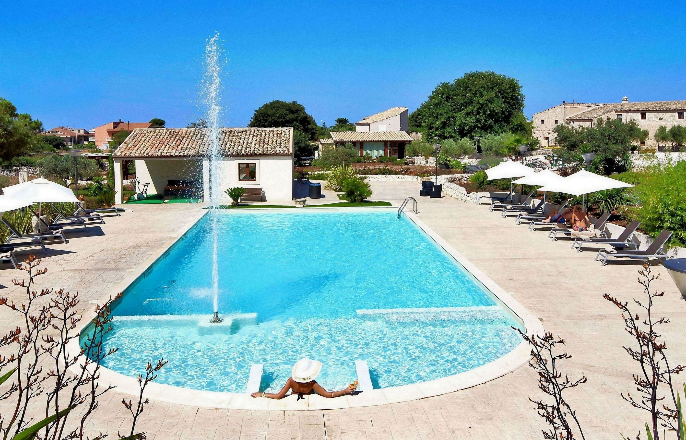 A swimming pool with a fountain, surrounded by lounge chairs and umbrellas, with buildings in the background.
