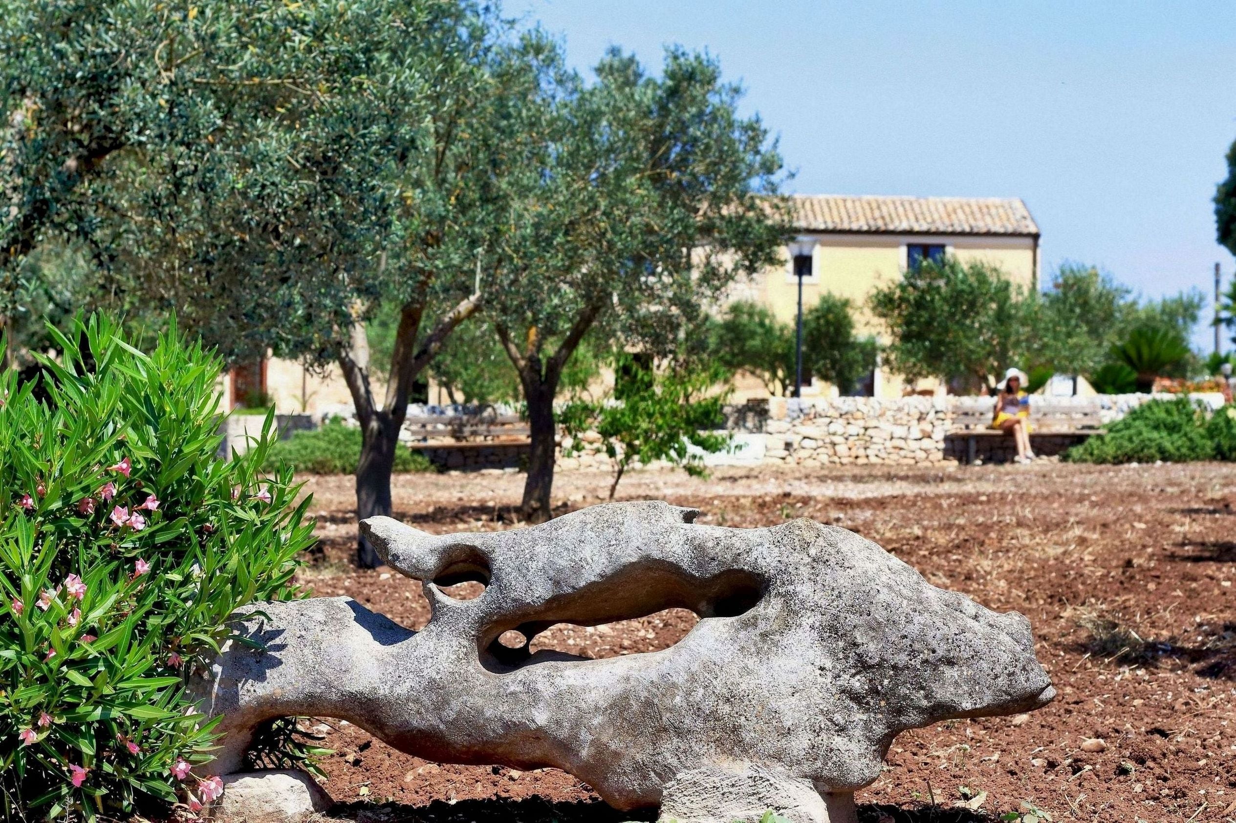 A stone sculpture in a garden with olive trees and a building in the background.