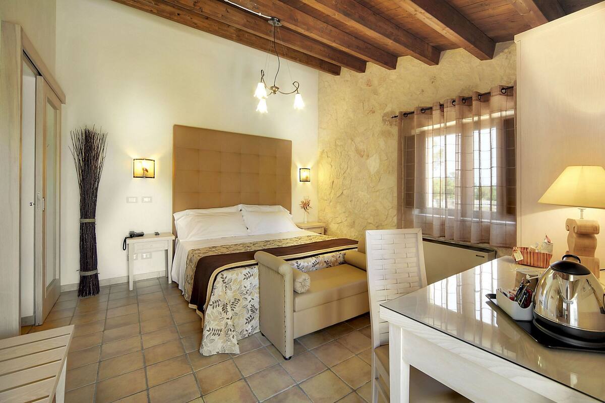 A bedroom with a bed, bedside table, lamp, and a view of the outside through a window.