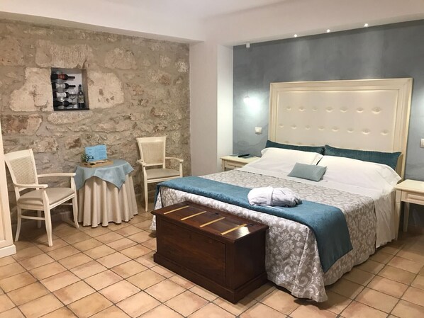 Un dormitorio con pared de piedra, una cama con colcha azul, un cofre de madera y una pequeña mesa con sillas.