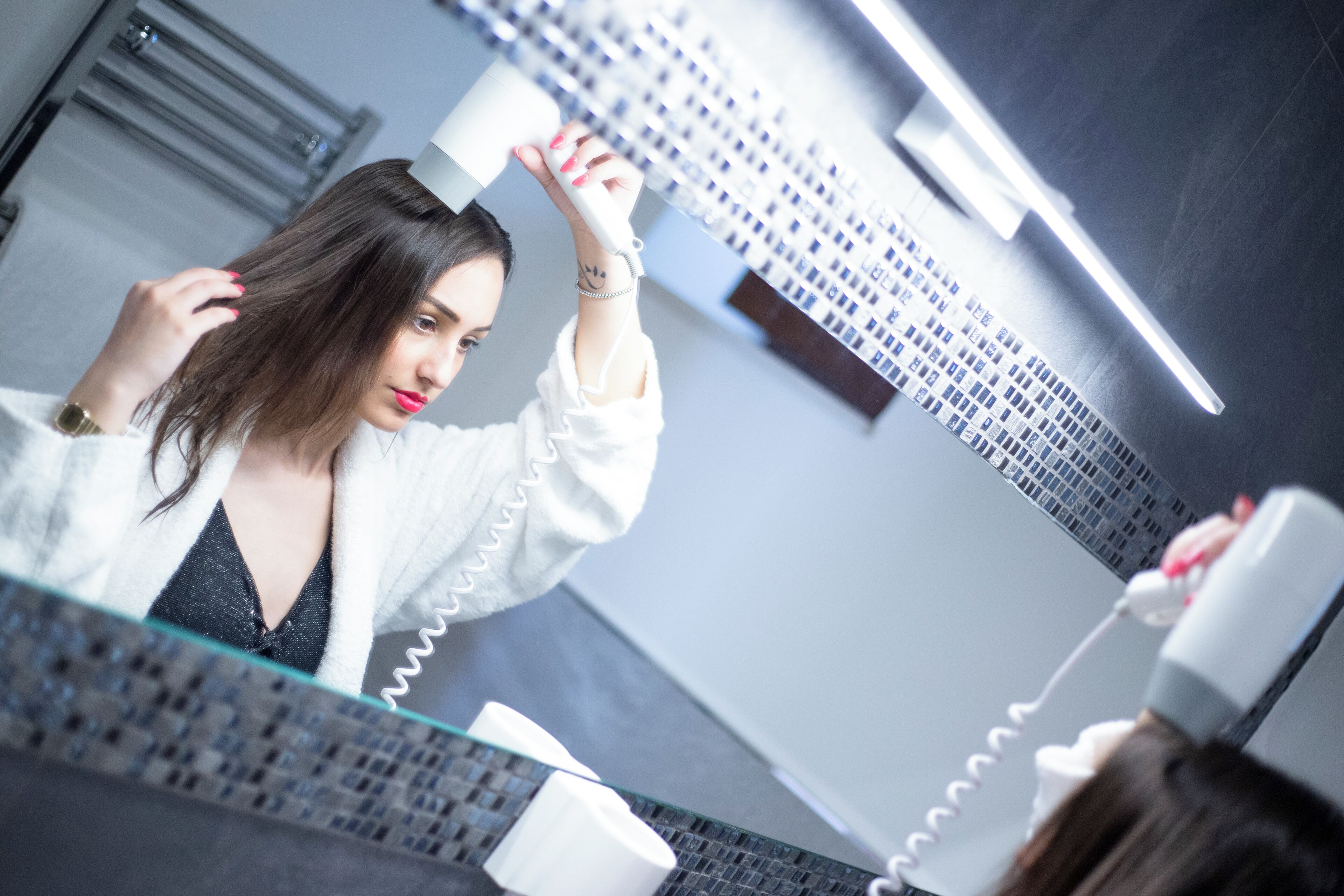 Una persona usando un secador de pelo en un baño con pared de azulejos y un espejo.
