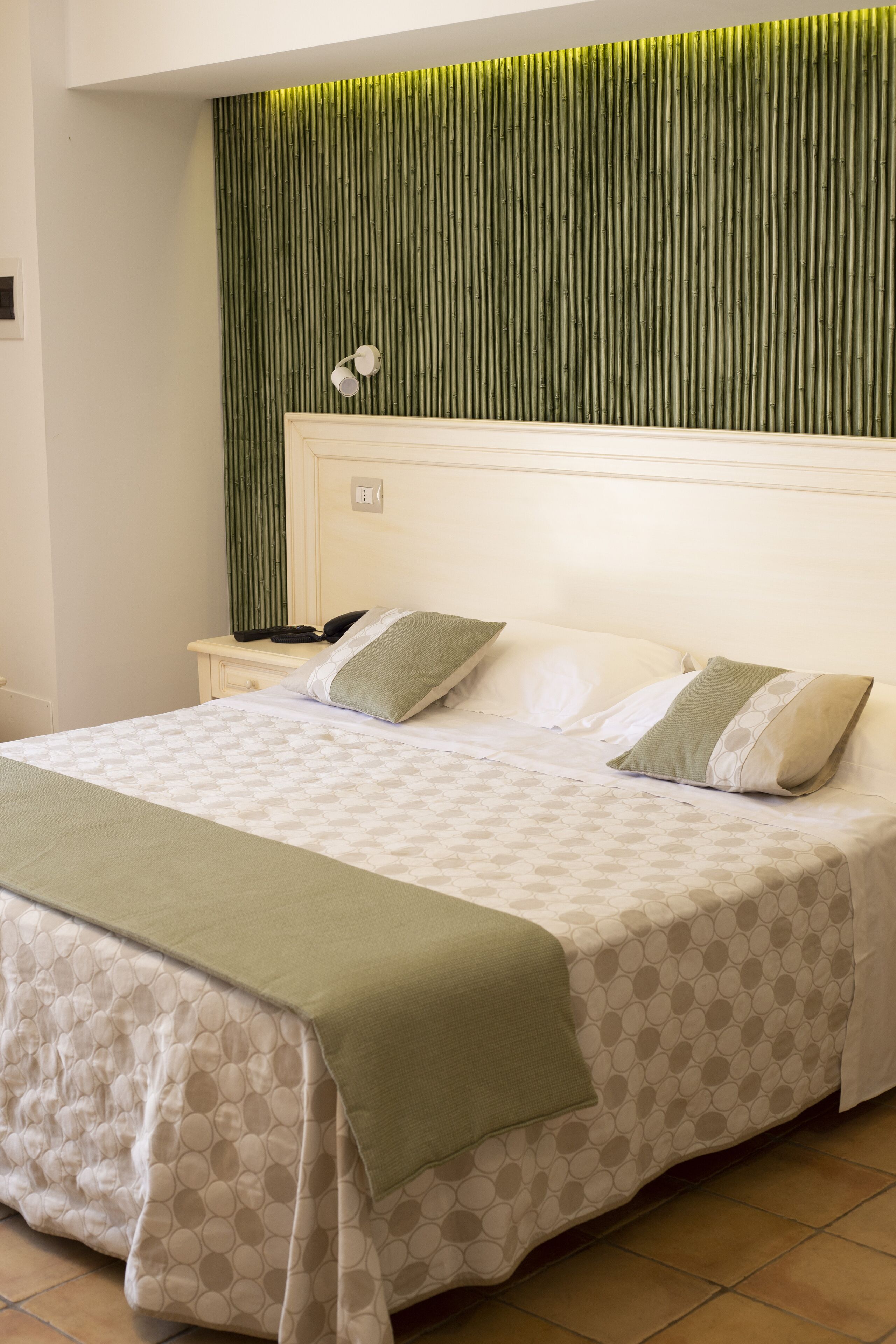 Una cama bien hecha con colcha y almohadones estampados, una mesita de noche con teléfono y lámpara, y una pared con paneles verticales que recuerdan a la caña de bambú.