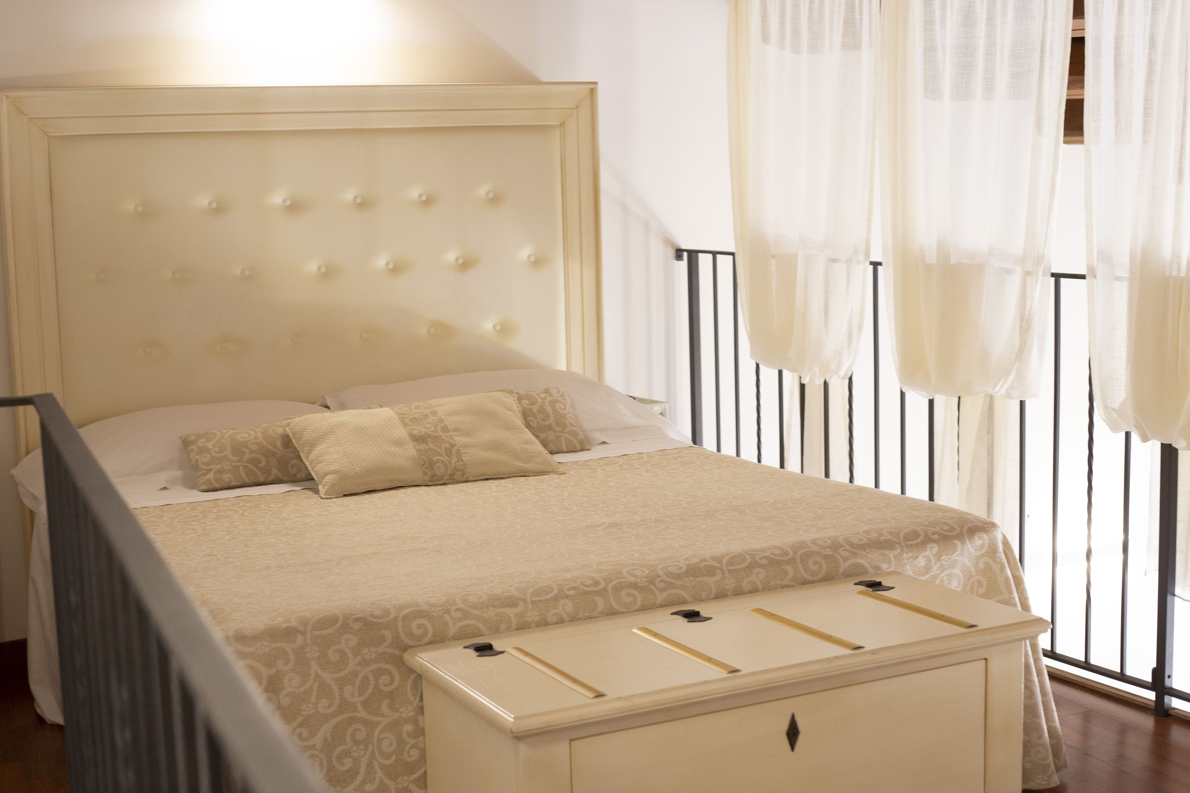 Una cama bien tendida con cabecera capitonada, una mesita de noche y una ventana con cortinas de gasa.