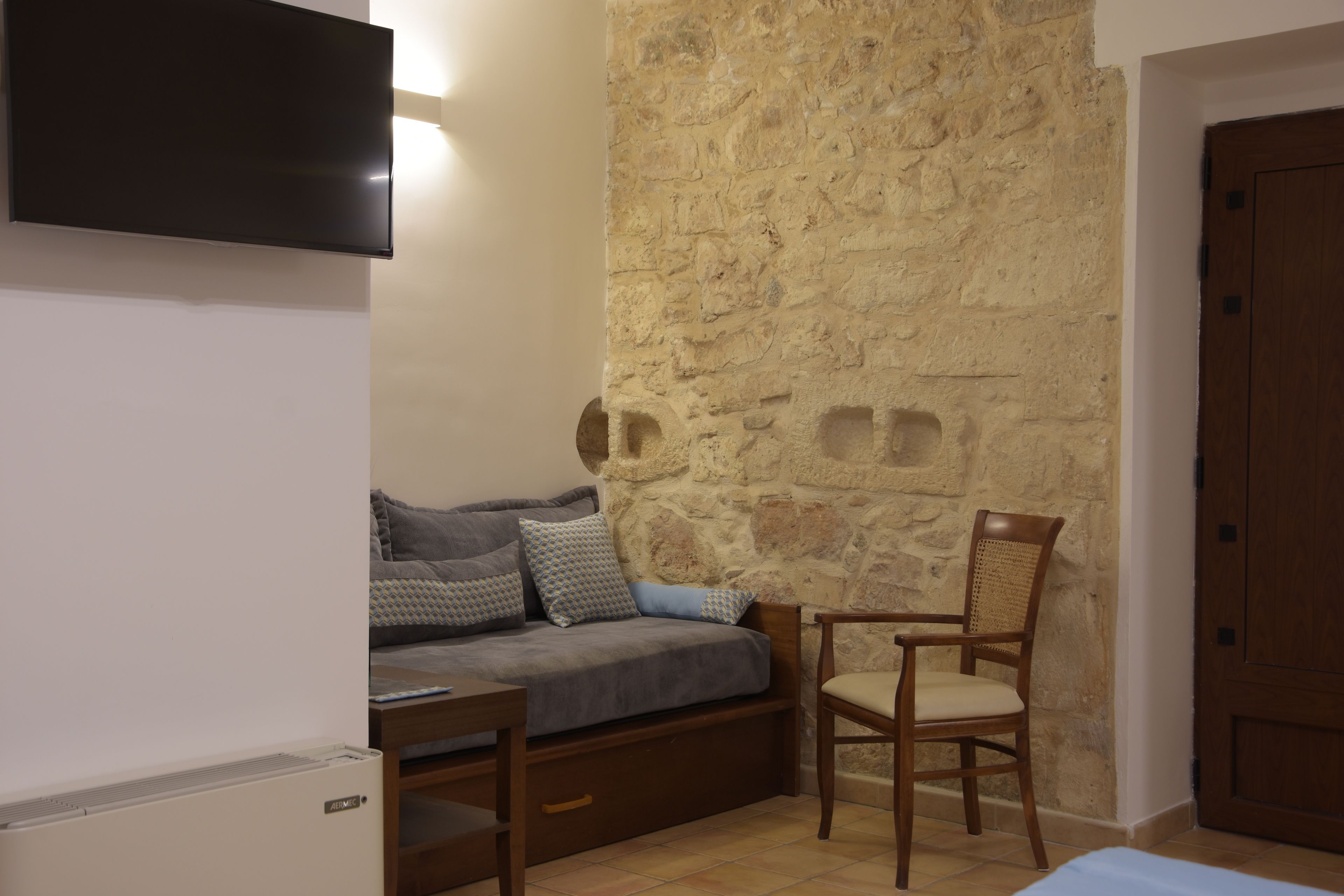 Een gezellige kamer met een stenen muur, een bank met kussens, een houten stoel, een aan de muur bevestigde televisie en een houten deur.