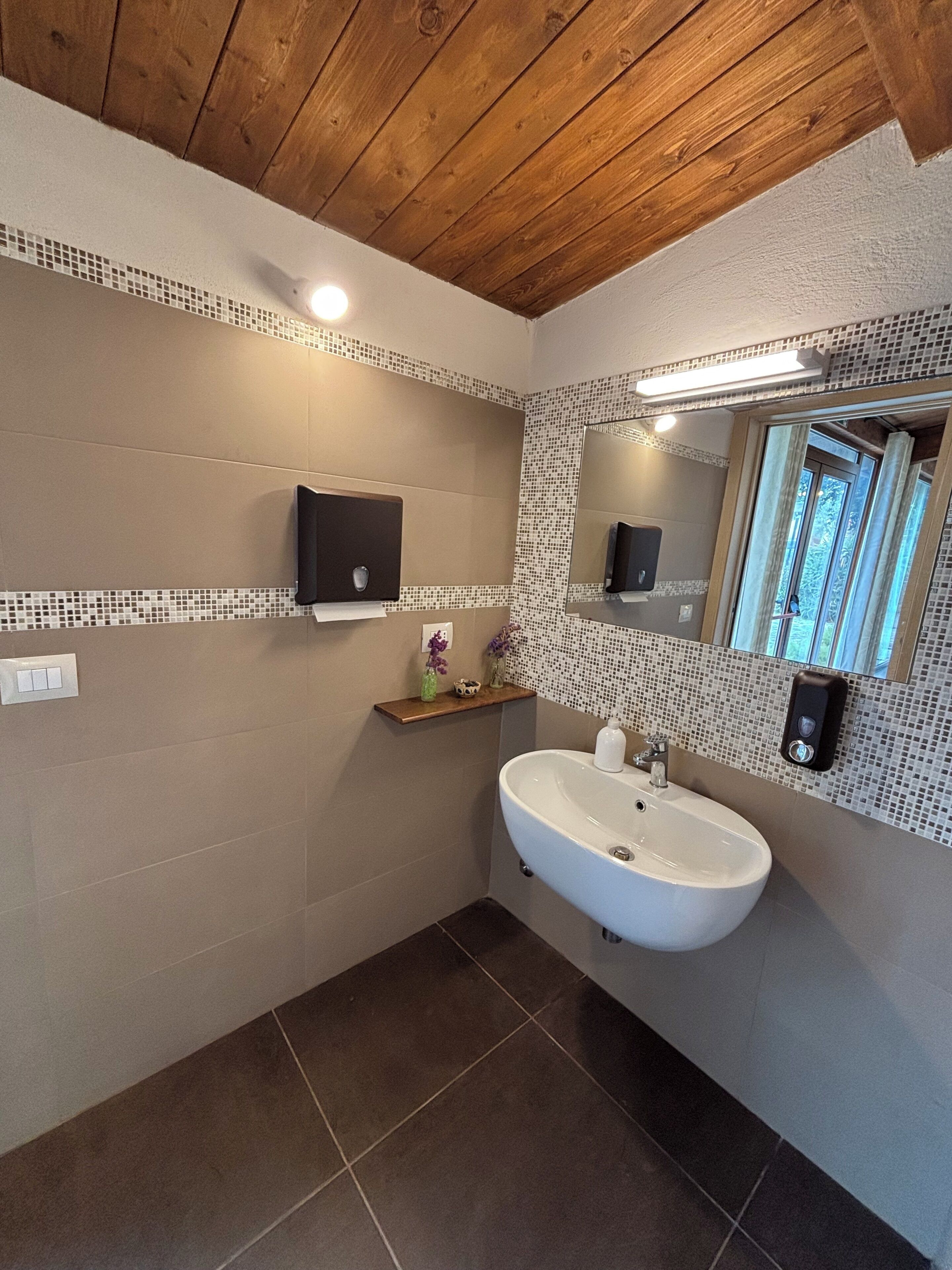 Un baño con techo de madera, un lavamanos blanco, un espejo y una estantería con artículos de aseo.