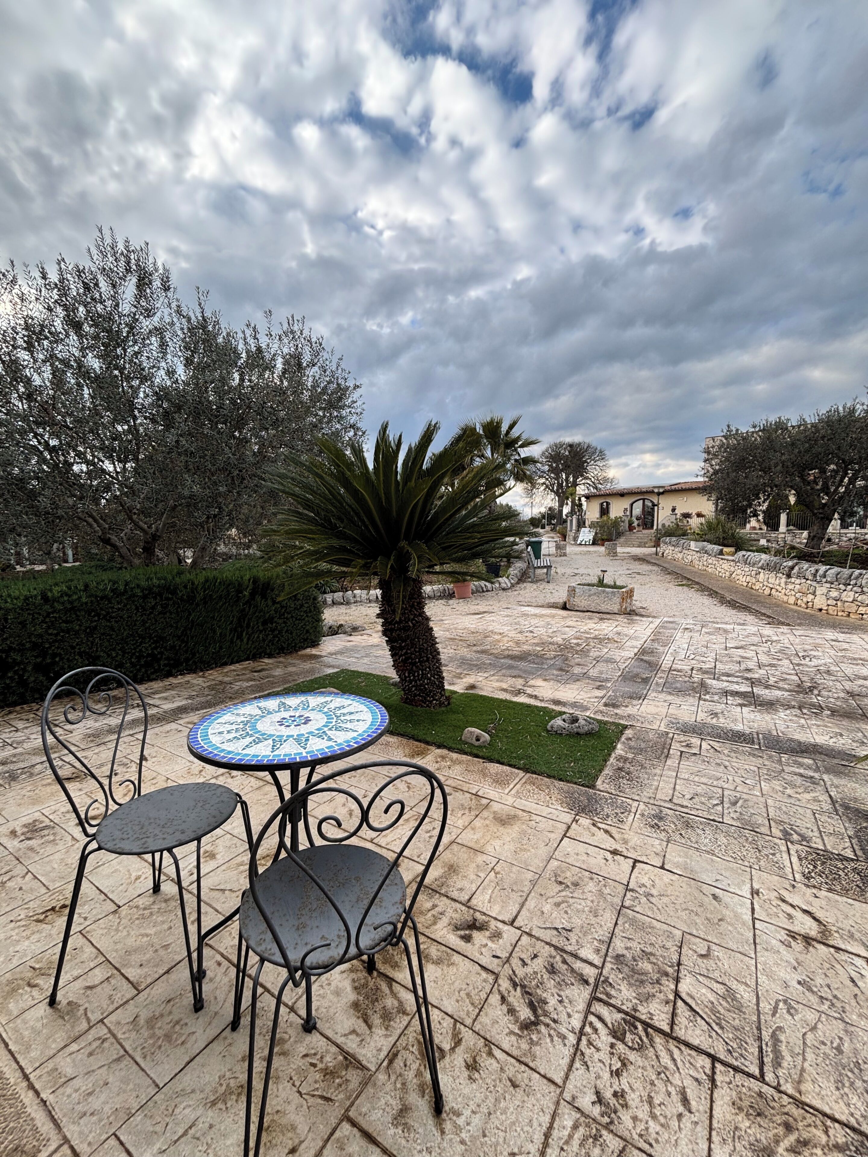 Un patio al aire libre con una mesa redonda y sillas, rodeado de baldosas de piedra y follaje.