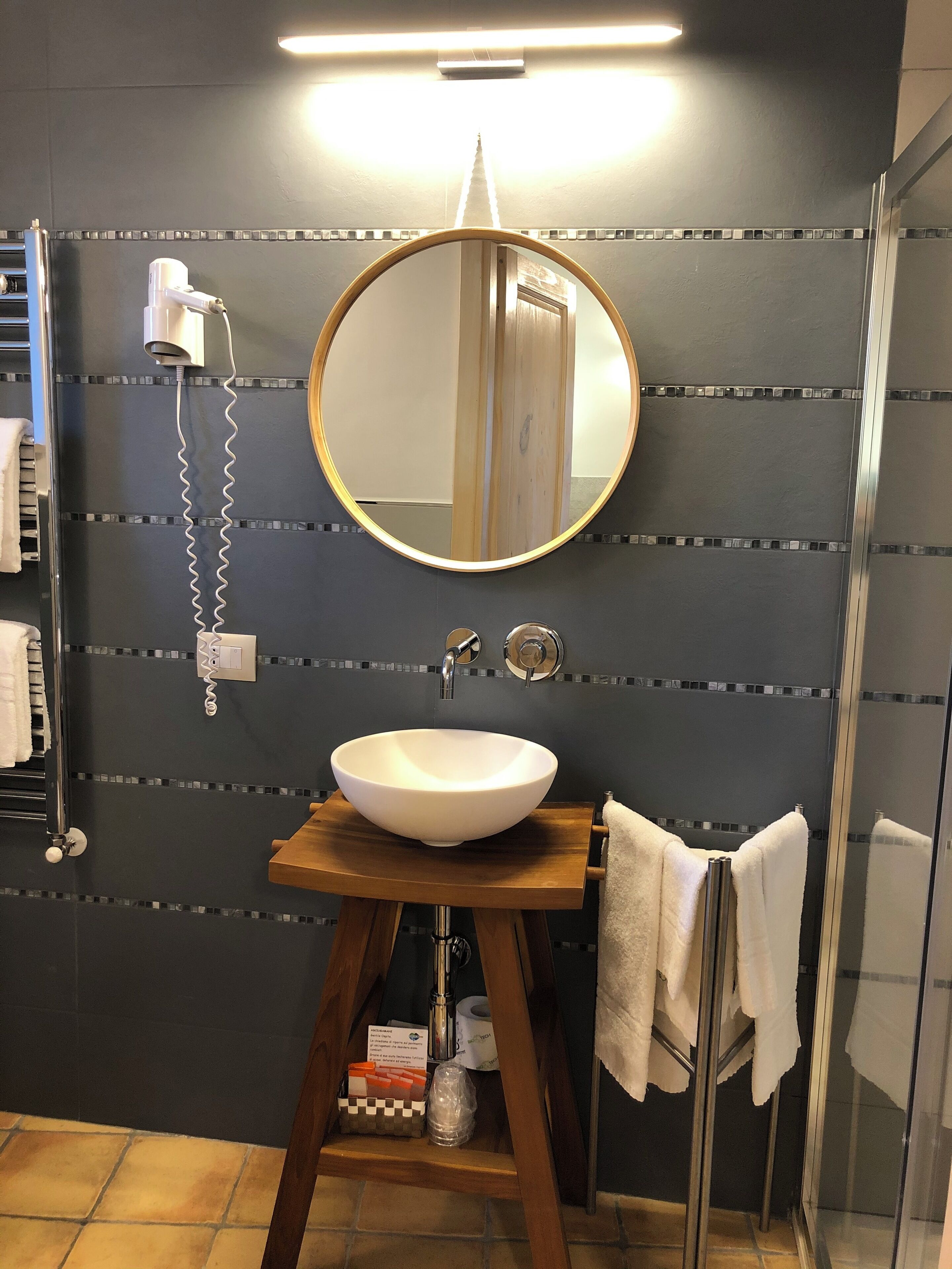 Une salle de bain avec un miroir rond, un lavabo blanc, un porte-serviettes, un sèche-cheveux et un espace douche.