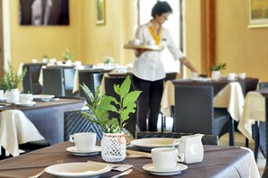 A dining area with tables set for service, a waiter in the background, and a decorative plant centerpiece. - Artemisia Resort (Ragusa)