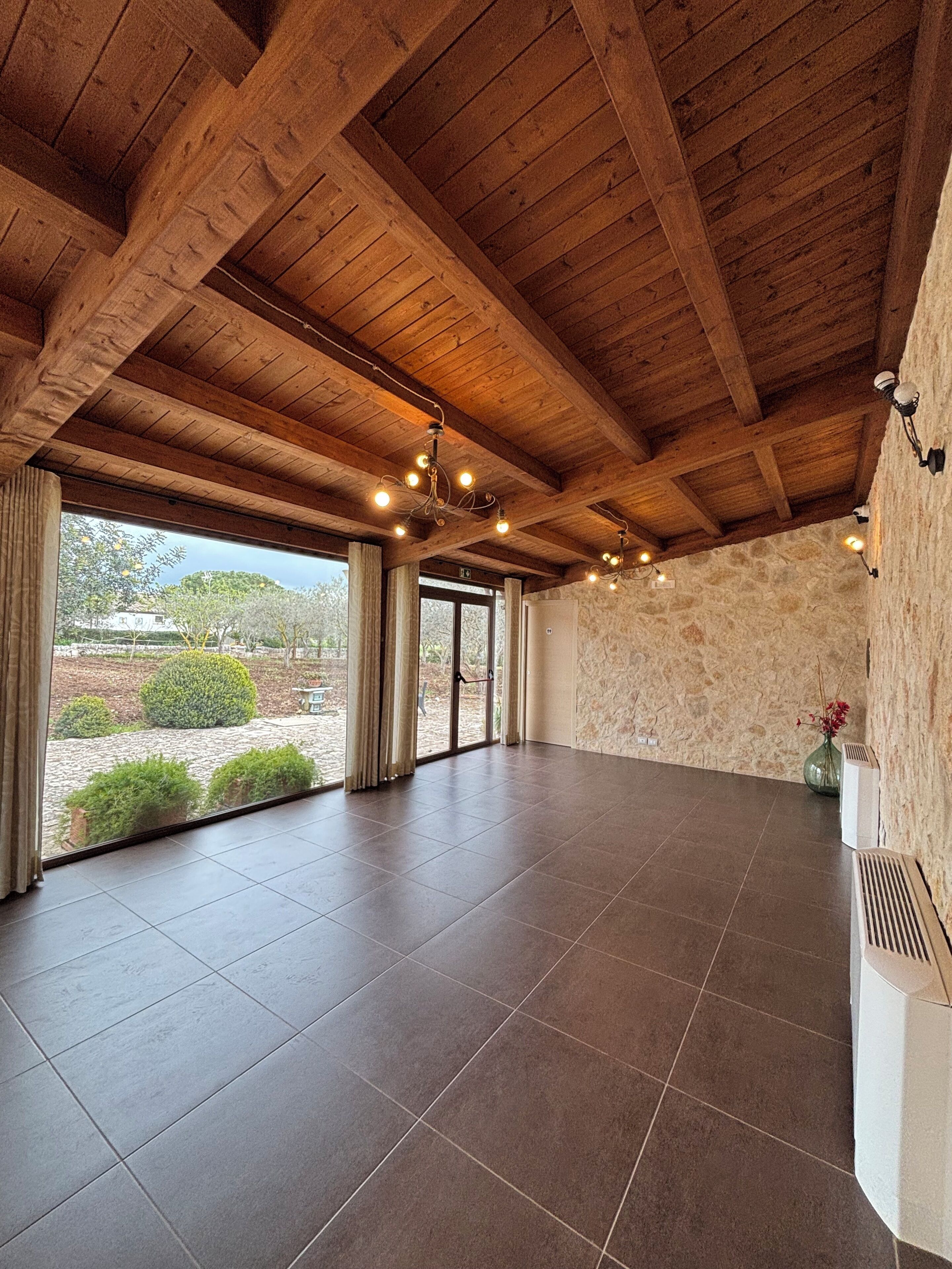 A spacious room with a high wooden ceiling, large windows, and a stone wall.