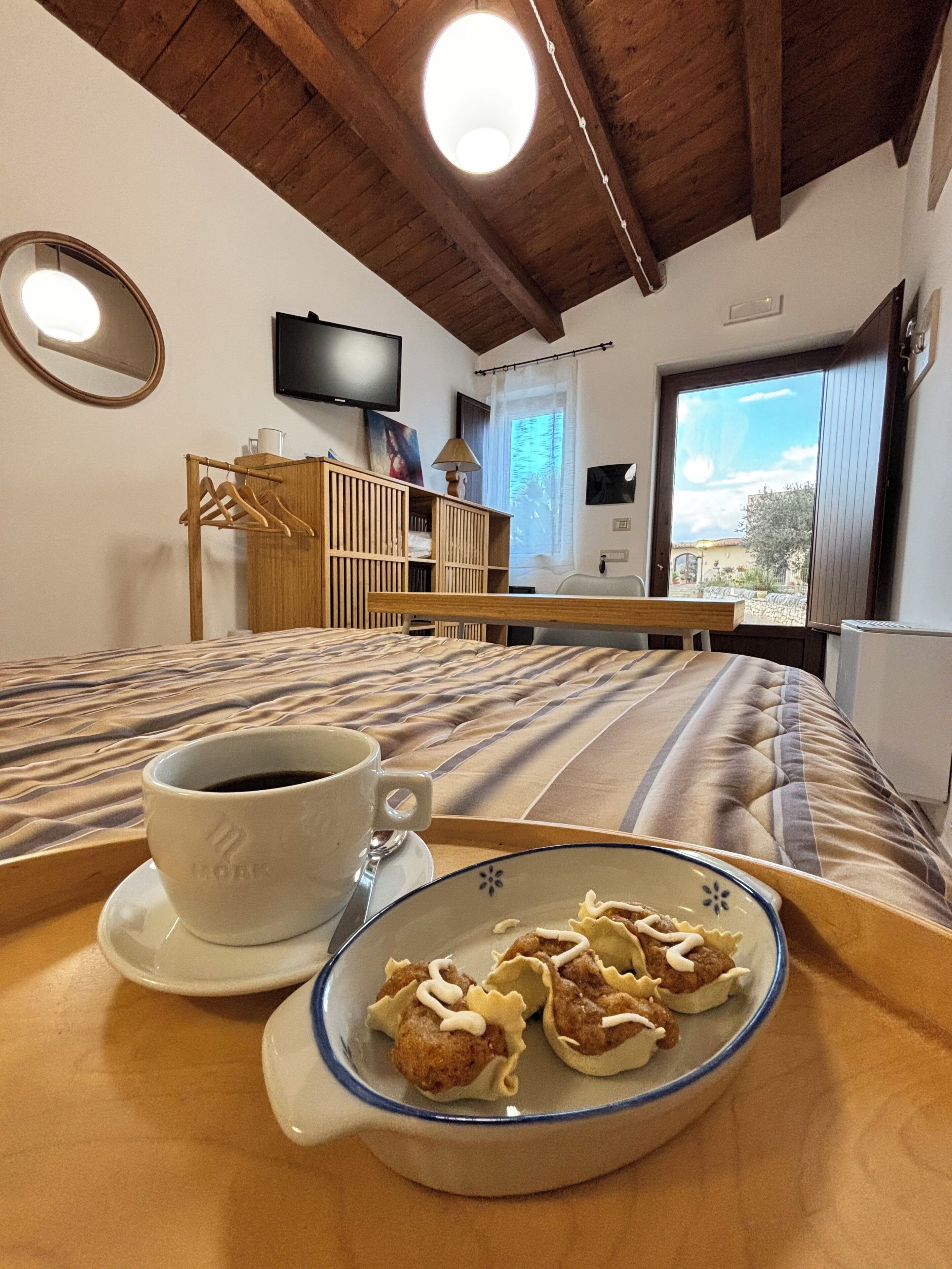Une chambre avec un lit, une table de chevet sur laquelle se trouvent une tasse de café et des viennoiseries, un téléviseur fixé au mur et une fenêtre donnant sur un beau paysage.