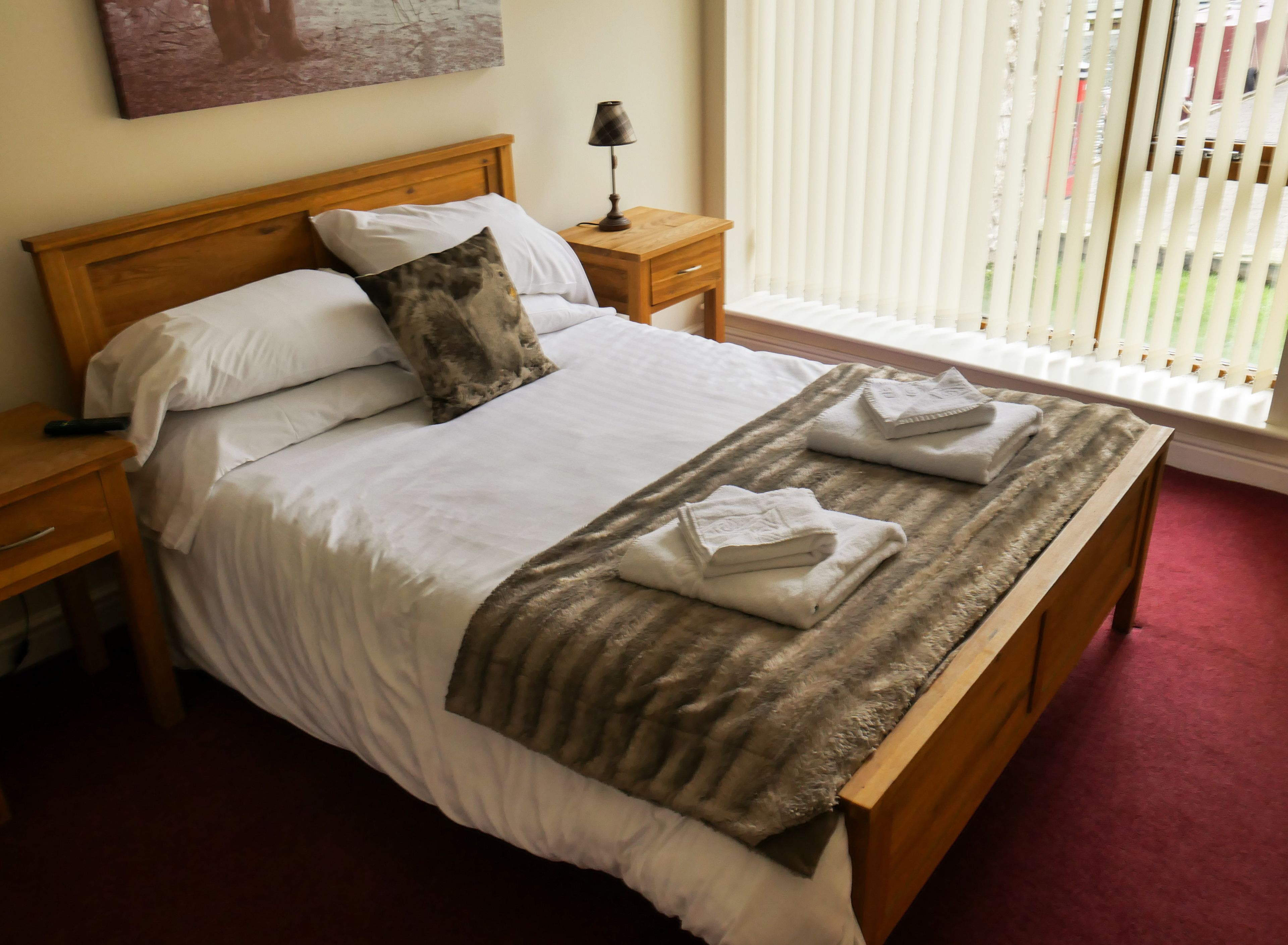 A neatly made bed with white linens, a brown throw blanket, and two folded towels on the footboard. There is a wooden nightstand with a lamp beside the bed, and a painting on the wall above it.