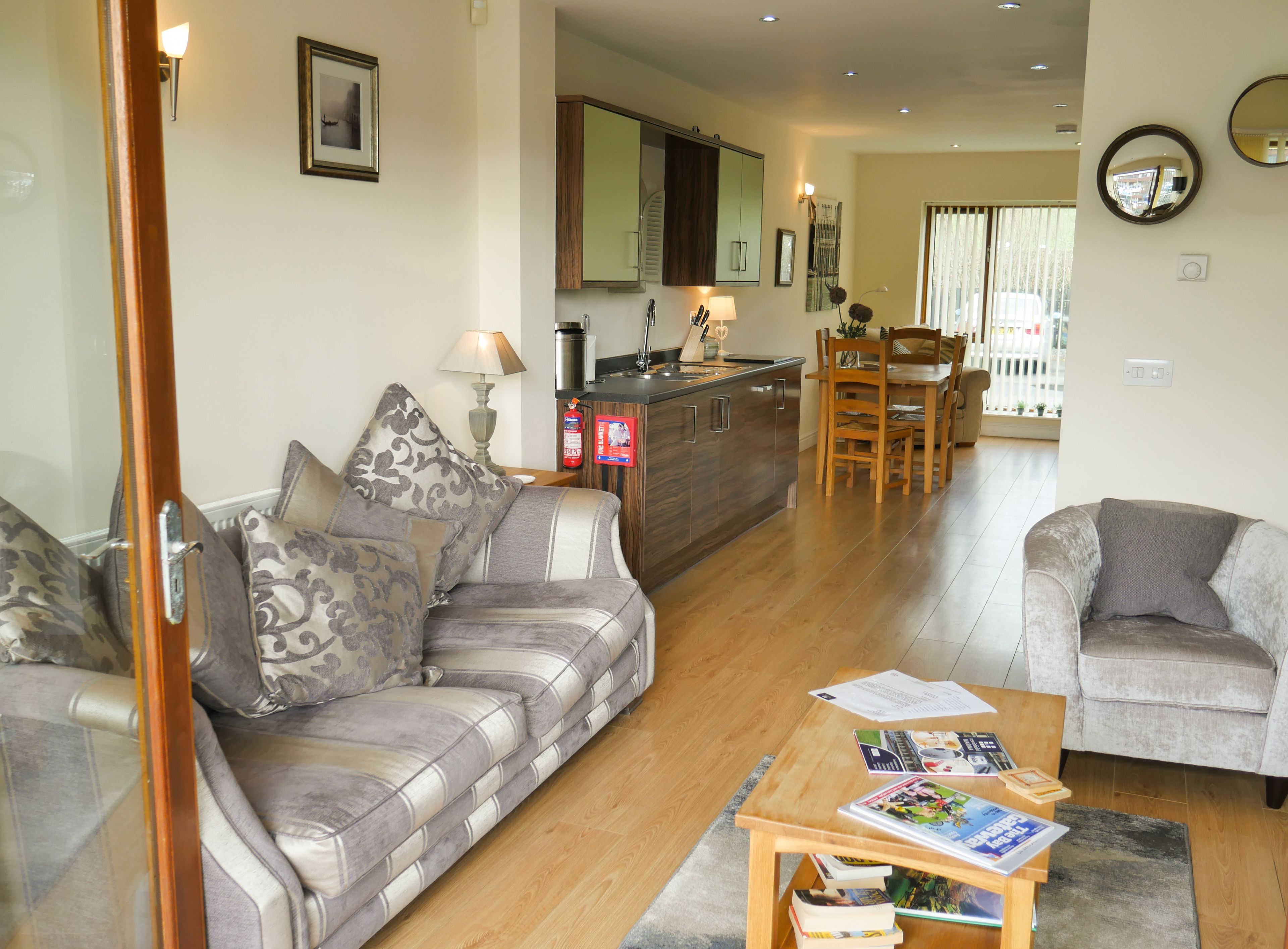 A living room with a sofa, armchair, and a coffee table with books. There is a kitchen area with a dining table and chairs.