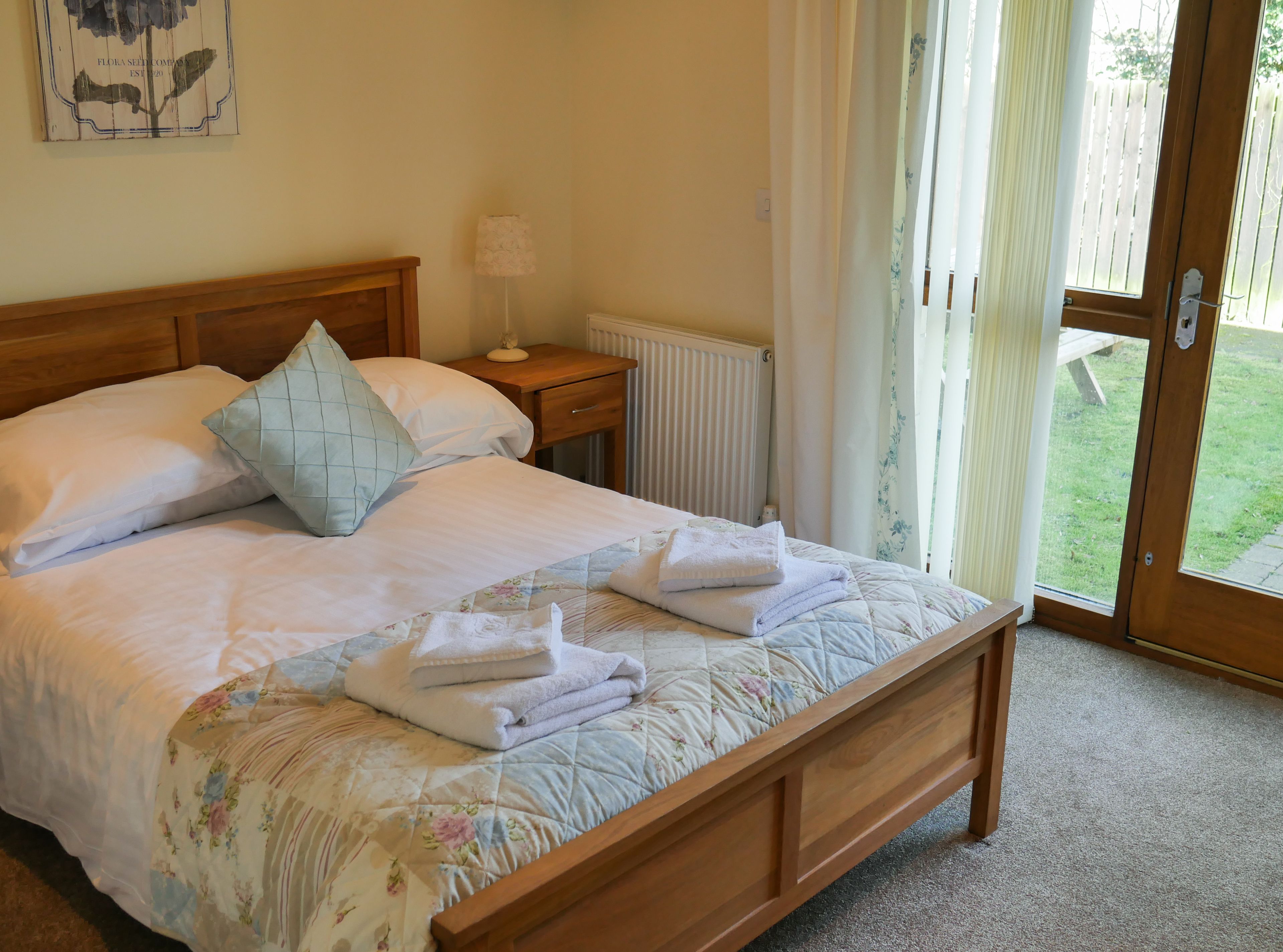 A neatly made bed with a floral duvet, a bedside lamp, and a view of a garden through an open door.