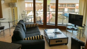 Una sala de estar con sofá, una mesa de centro de vidrio, televisión y vista a un canal con barcos.