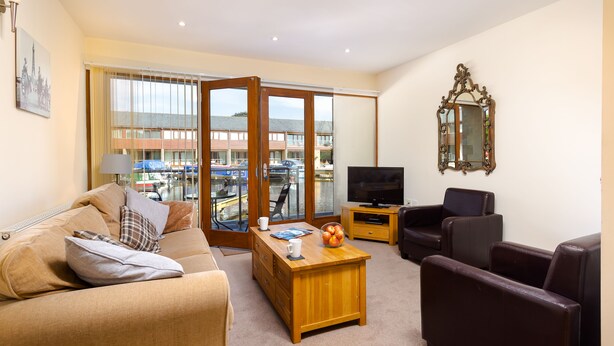 Sala de estar con sofá, televisión, mesa de centro de madera y vista a barcos a través de un ventanal.