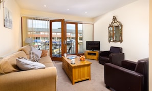 A living room with a sofa, a TV, a wooden coffee table, and a view of boats through a glass door.