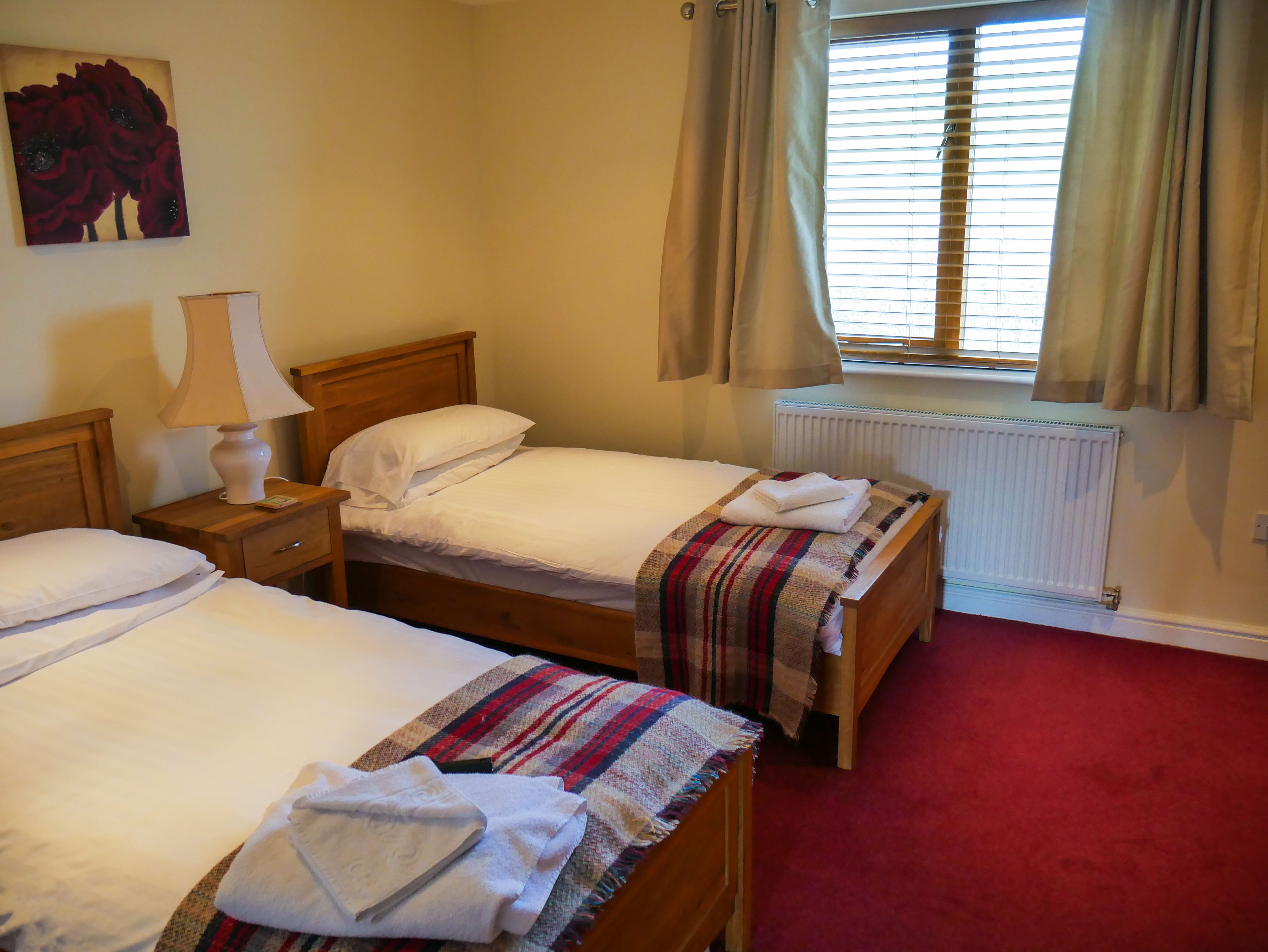A hotel room with two single beds, a wooden headboard, a bedside table with a lamp, a radiator, and a window with curtains.