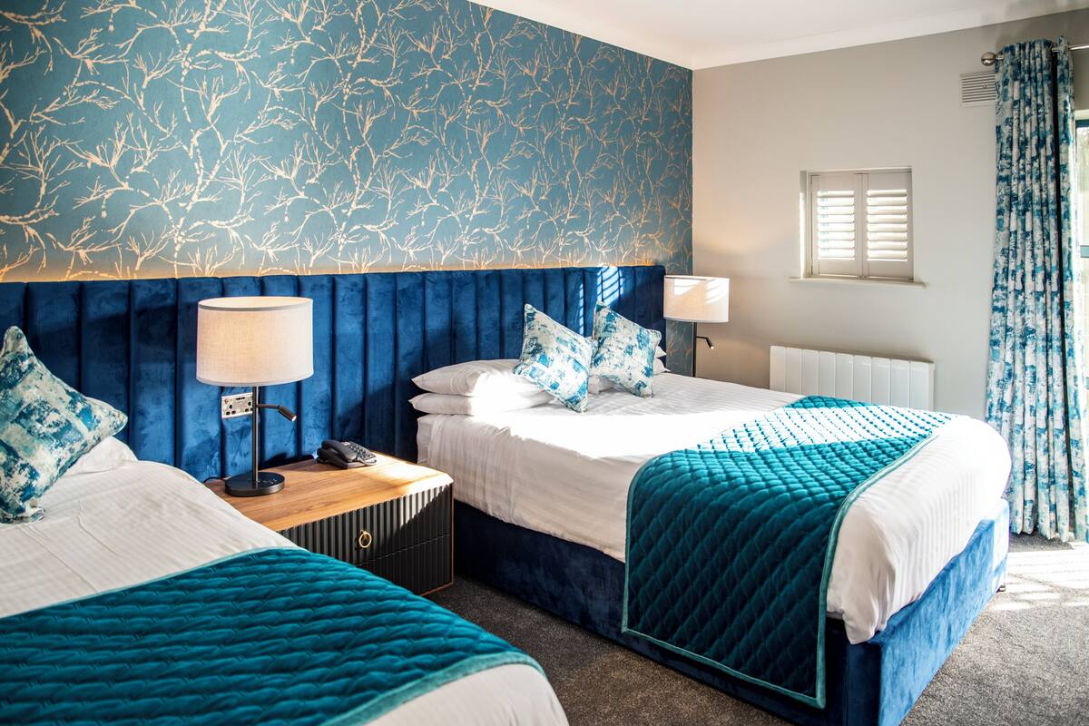 A hotel room with a large bed, a smaller bed, a nightstand with a lamp, a telephone, and a window with shutters.