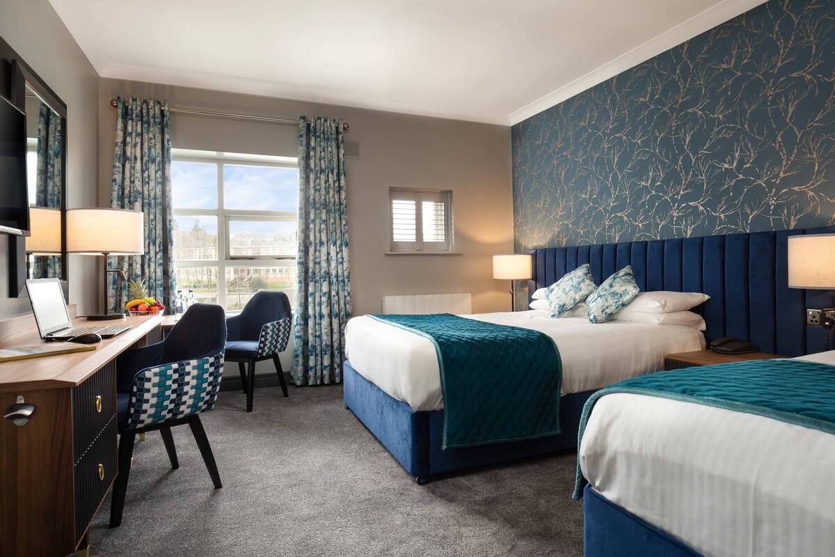 A hotel room with two beds, a desk with a laptop, a chair, a desk lamp, and a view of the outdoors.