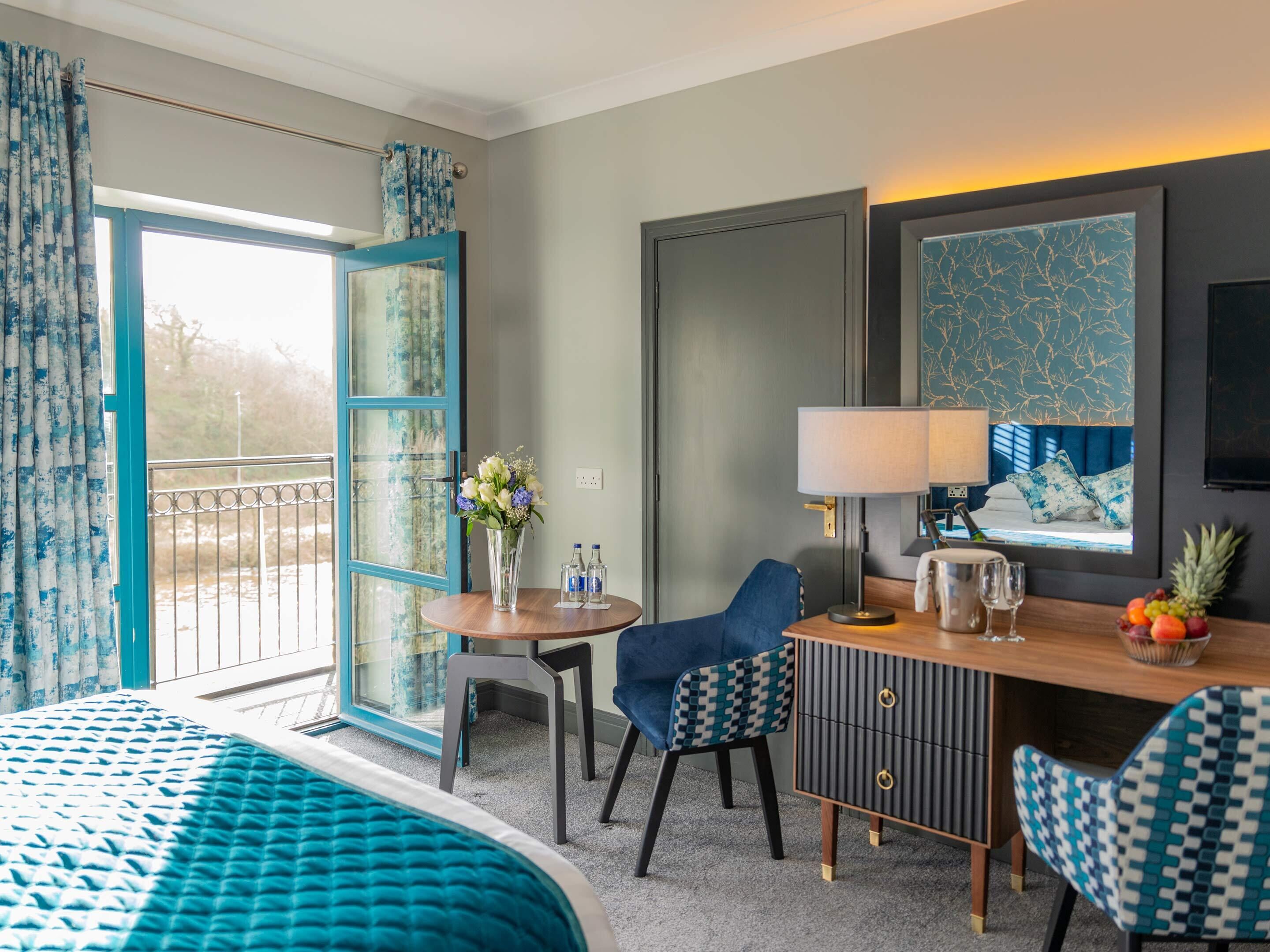 A hotel room with a bed, a desk with a chair, a dining table with a chair, a large window with curtains, and a view of the outdoors.