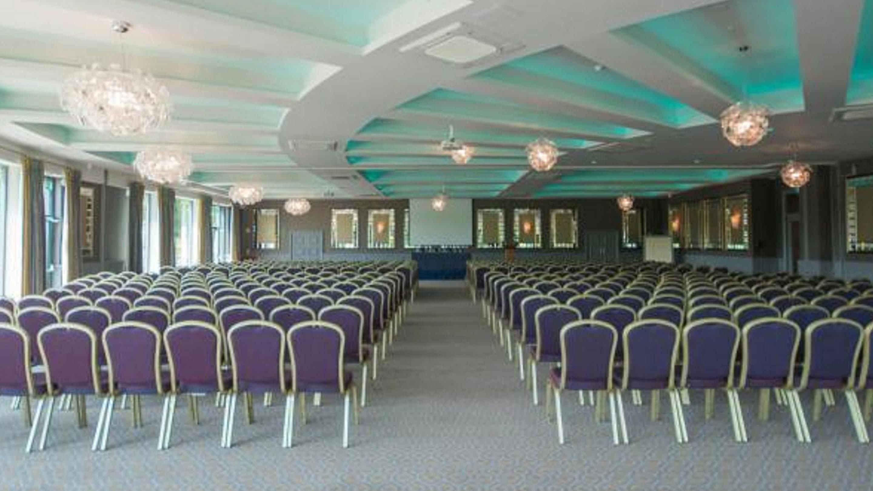 A conference room with rows of chairs, large windows, and decorative ceiling lights.