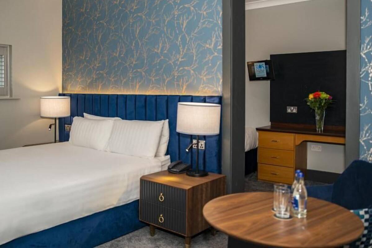 A hotel room with a large bed, two bedside lamps, a small wooden nightstand, a round table with a vase and water bottles, a TV on a wooden cabinet, and a patterned wall.