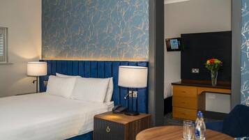 A hotel room with a large bed, two bedside lamps, a small wooden nightstand, a round table with a vase and water bottles, a TV on a wooden cabinet, and a patterned wall.