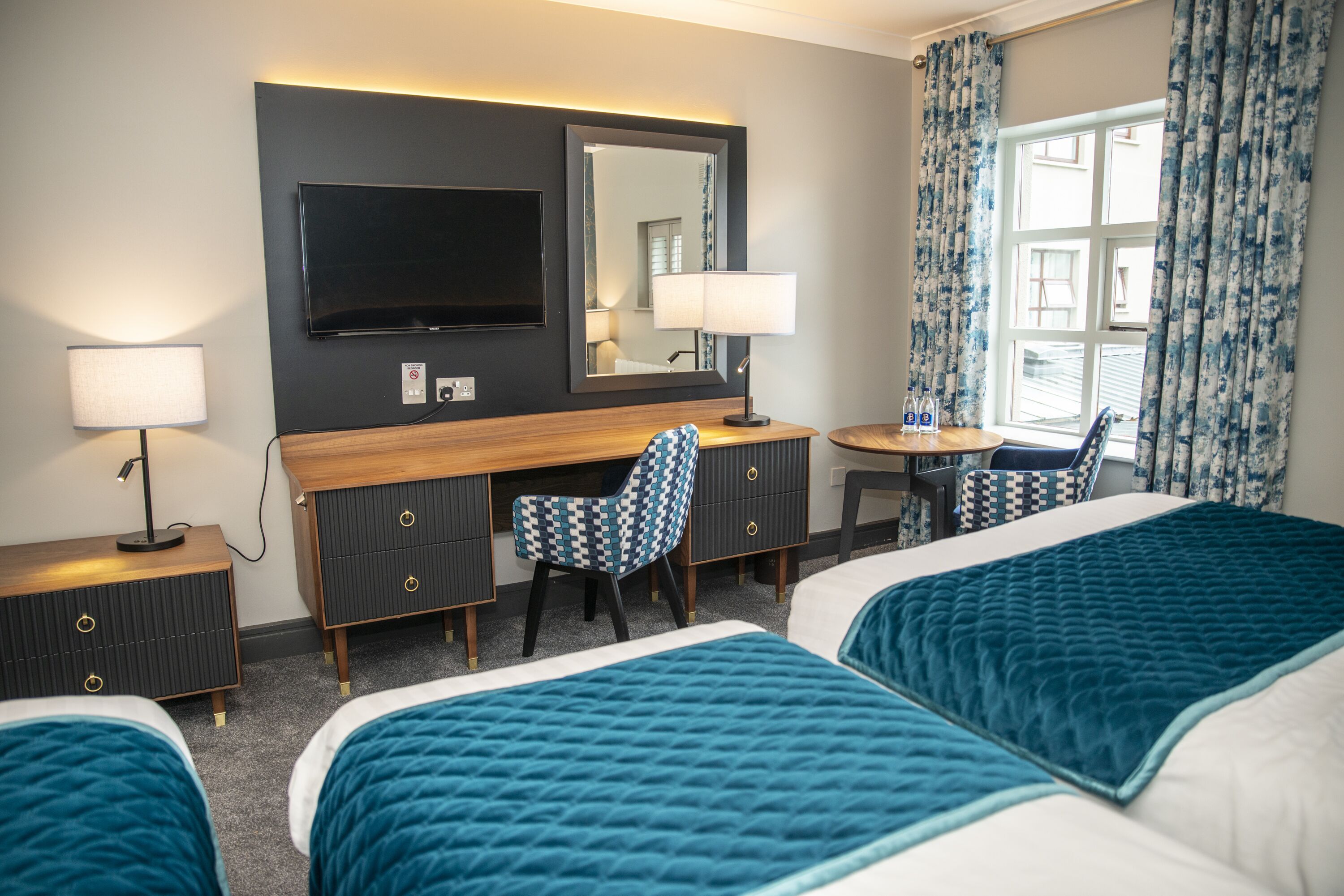 A hotel room with two beds, a desk, a chair, a TV, and a window with curtains.