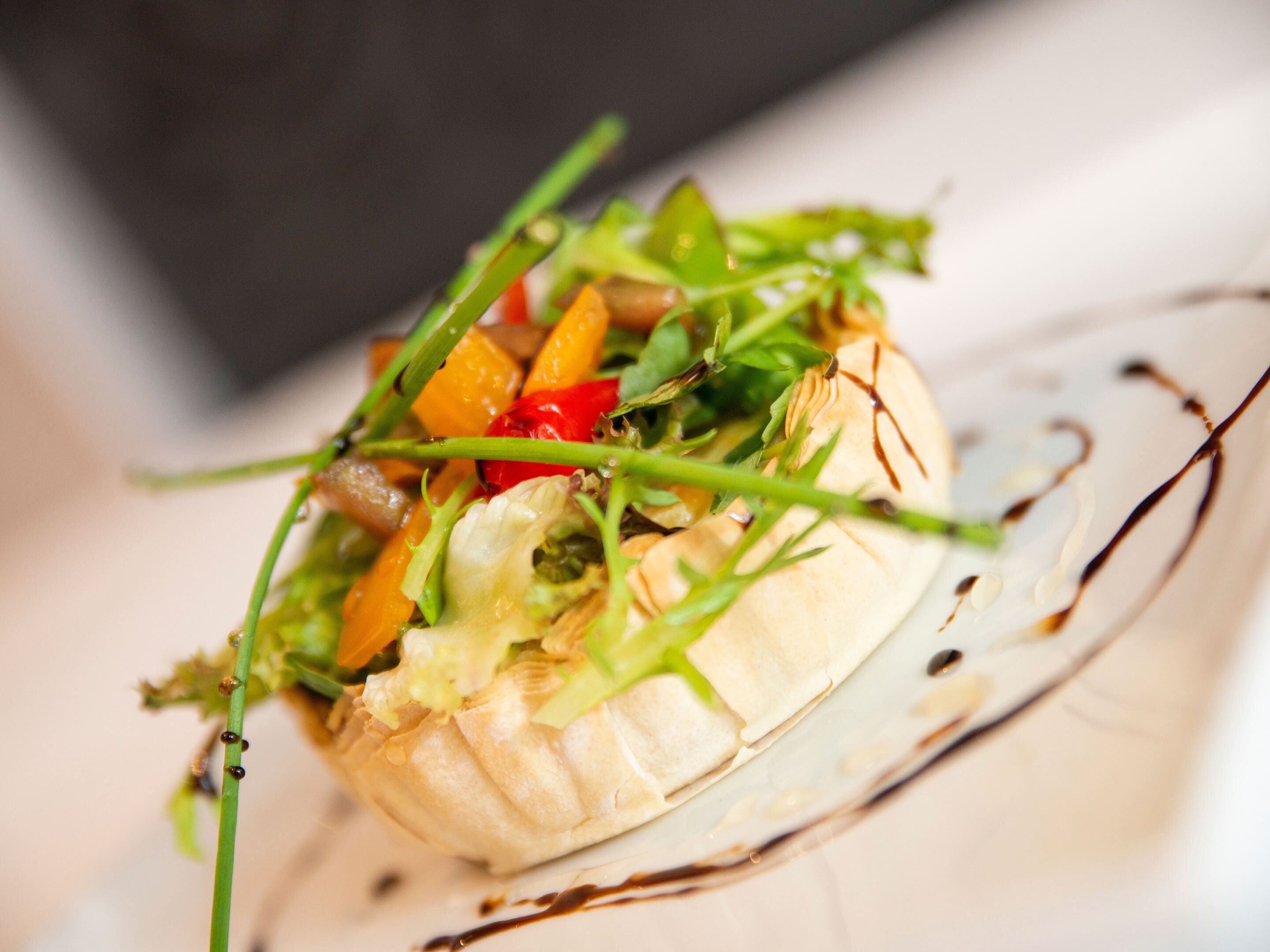 Une vue rapprochée d’un plat gastronomique composé de divers légumes et de différentes sortes de verdure, servi sur une assiette en verre transparent et agrémenté d’un filet de sauce.