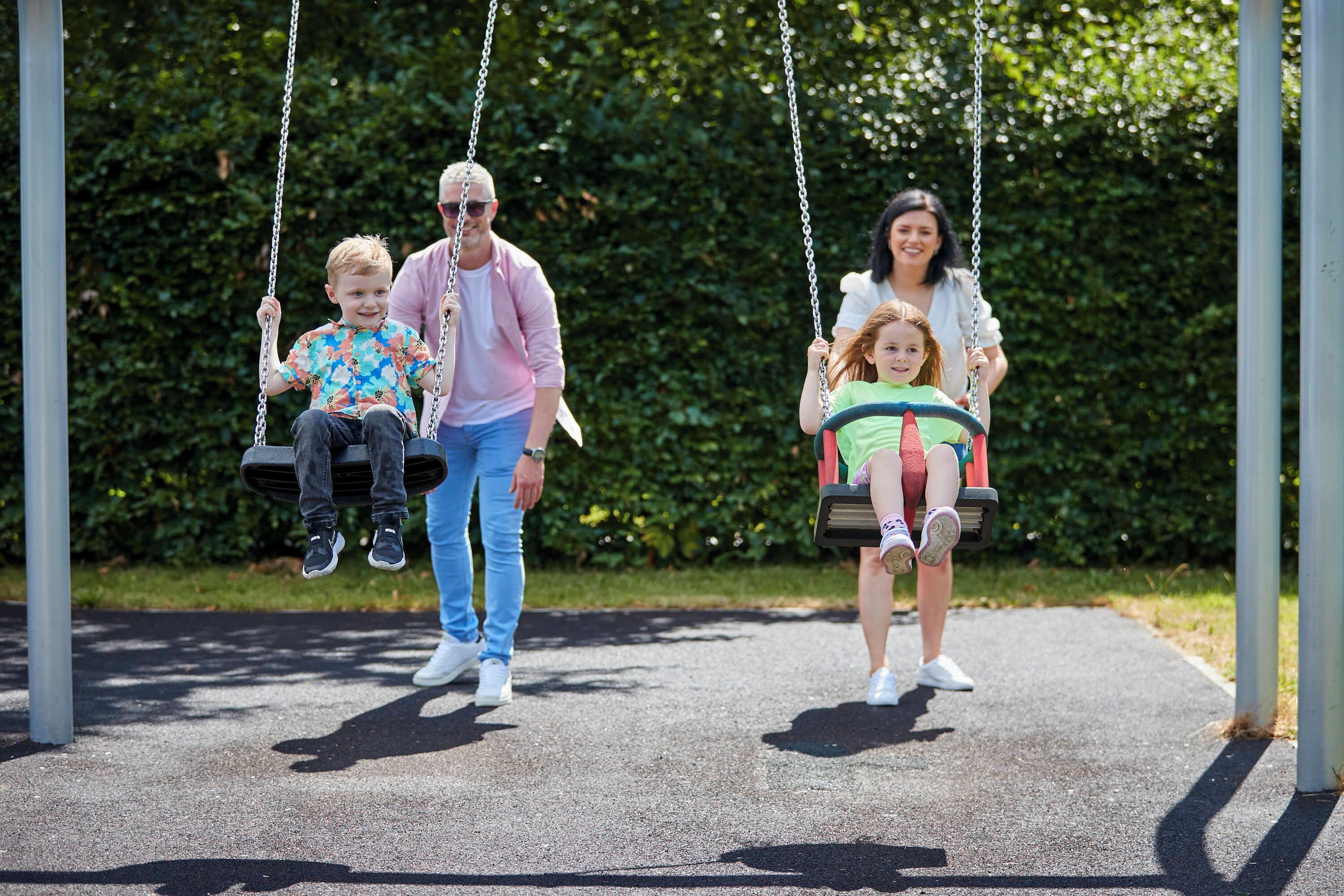 Eine Familie schwingt sich auf Schaukeln in einem Park mit einem befestigten Weg und Grün im Hintergrund.