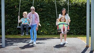 Eine Familie schwingt sich auf Schaukeln in einem Park mit einem befestigten Weg und Grün im Hintergrund.