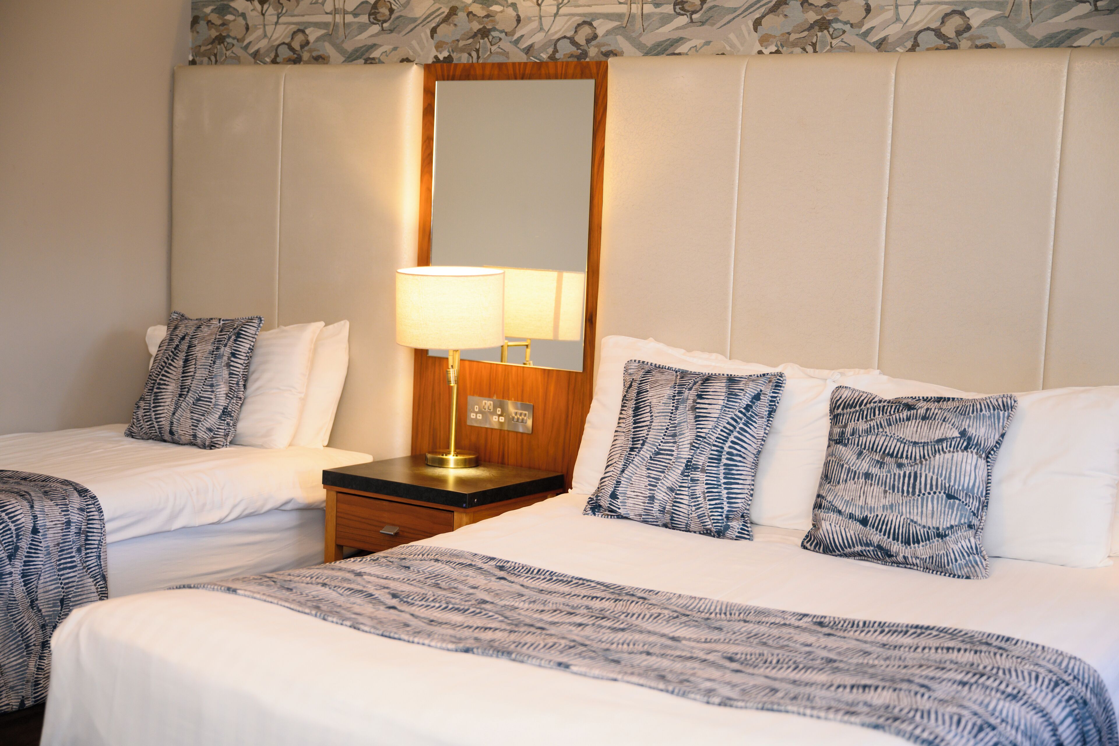 A hotel room with two beds, a nightstand with a lamp, and a large mirror.