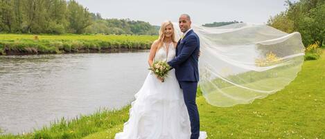 Un couple se tient au bord d’une rivière, avec un champ herbeux et des arbres en arrière-plan.