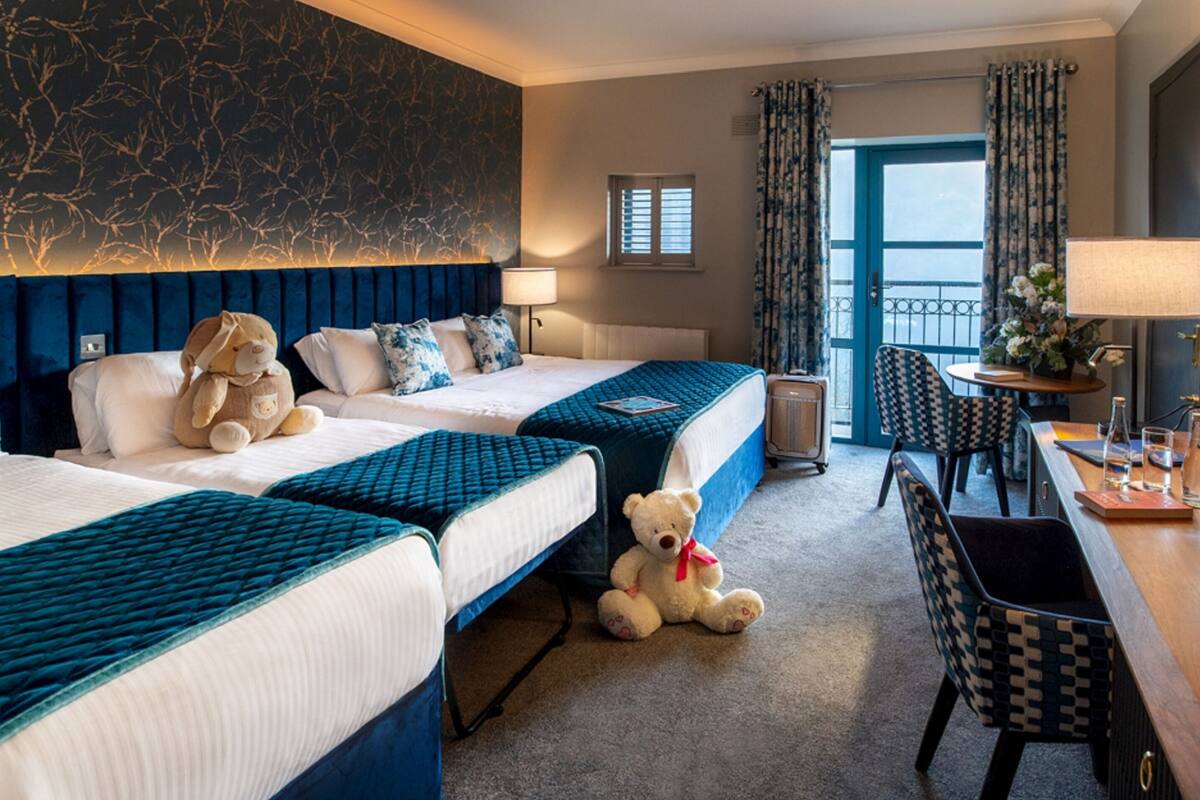 A hotel room with two beds, a desk, a chair, a teddy bear, and a window with curtains.