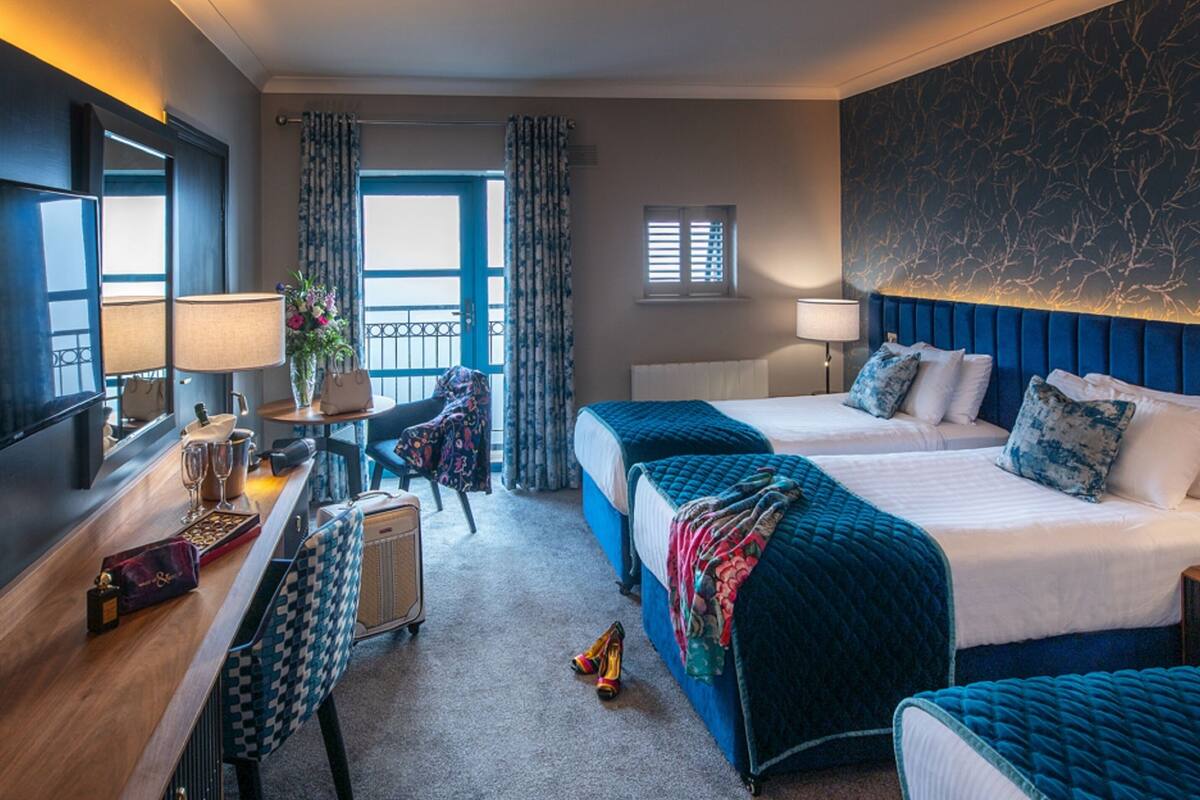 A hotel room with two beds, a desk with a lamp, a chair, a small table with a vase, and a window with curtains.