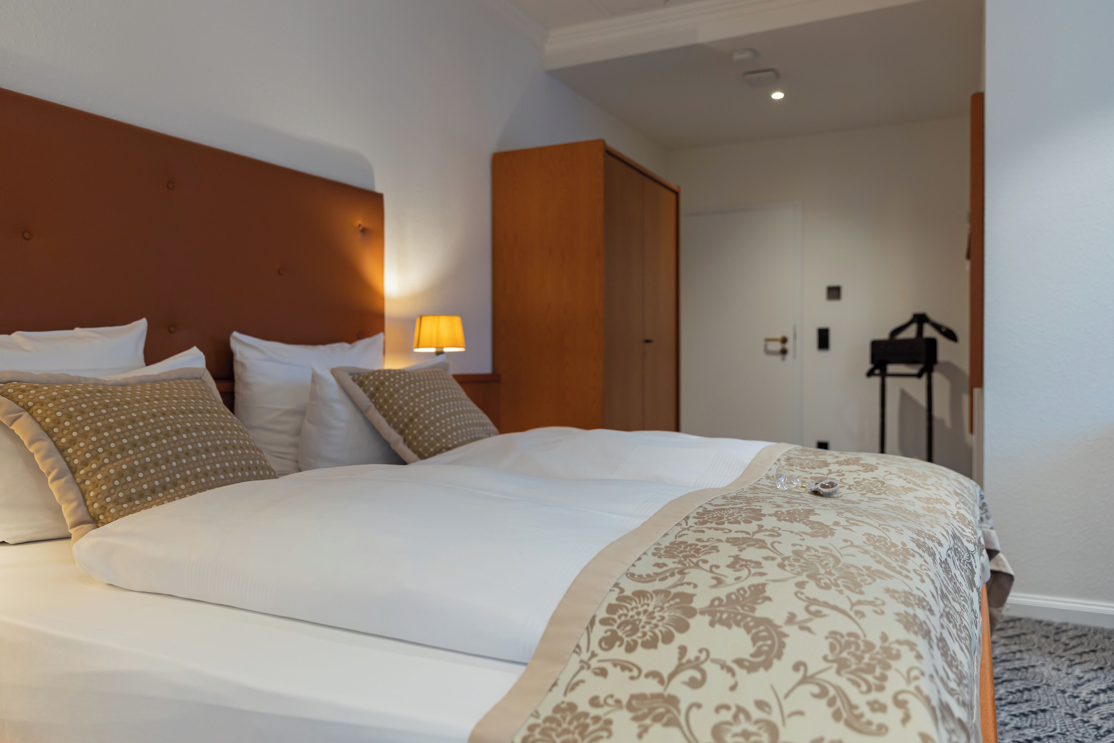 A neatly made bed with a patterned bedspread and pillows, a wooden headboard, and a lamp on a bedside table.