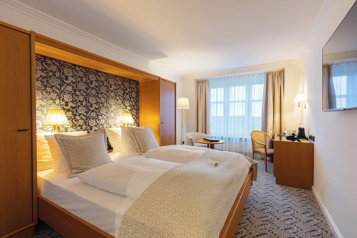 A hotel room with a large bed, two bedside lamps, a desk with a chair, and a large window with curtains.