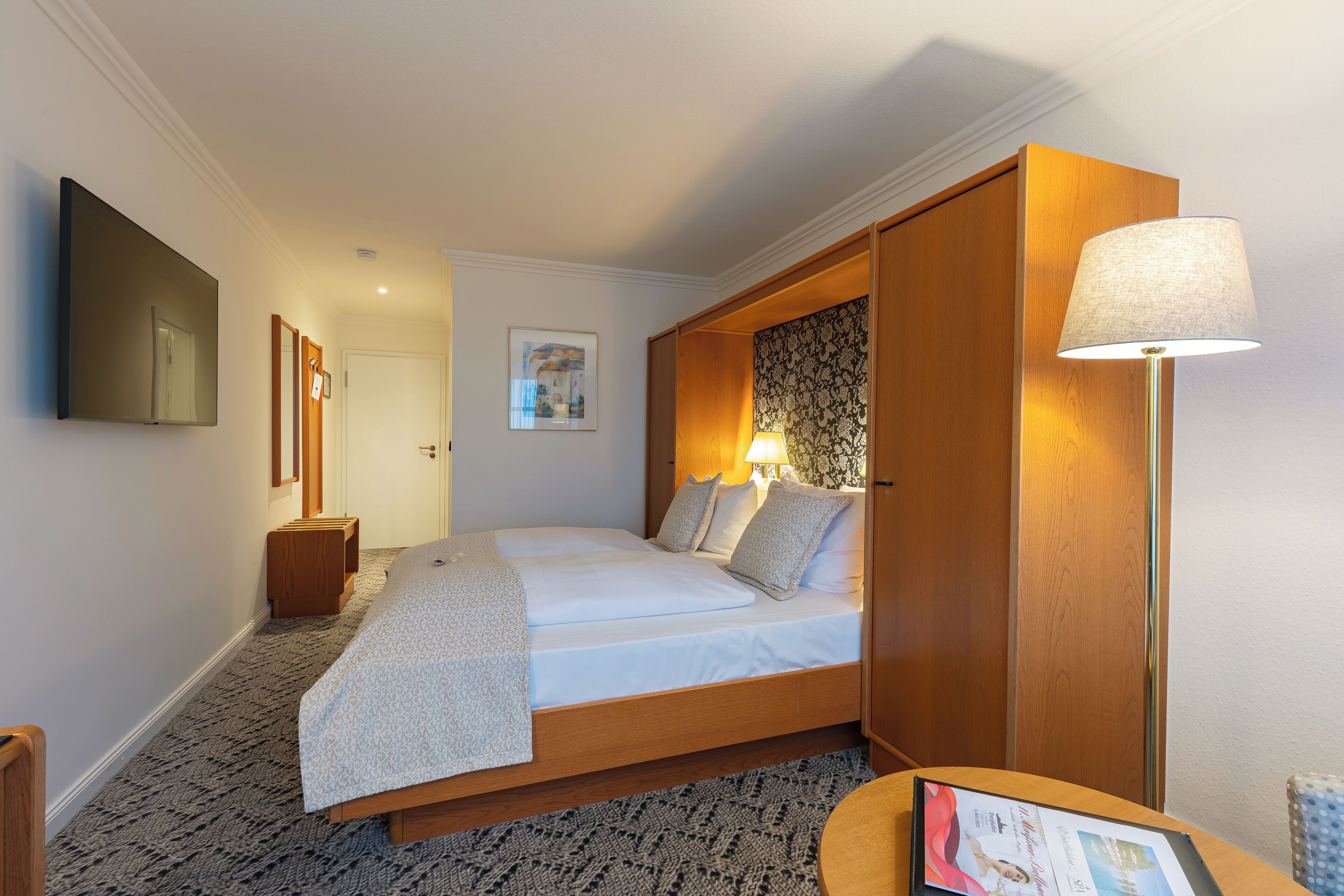 A bedroom with a bed, bedside lamp, and a flat-screen TV mounted on the wall.